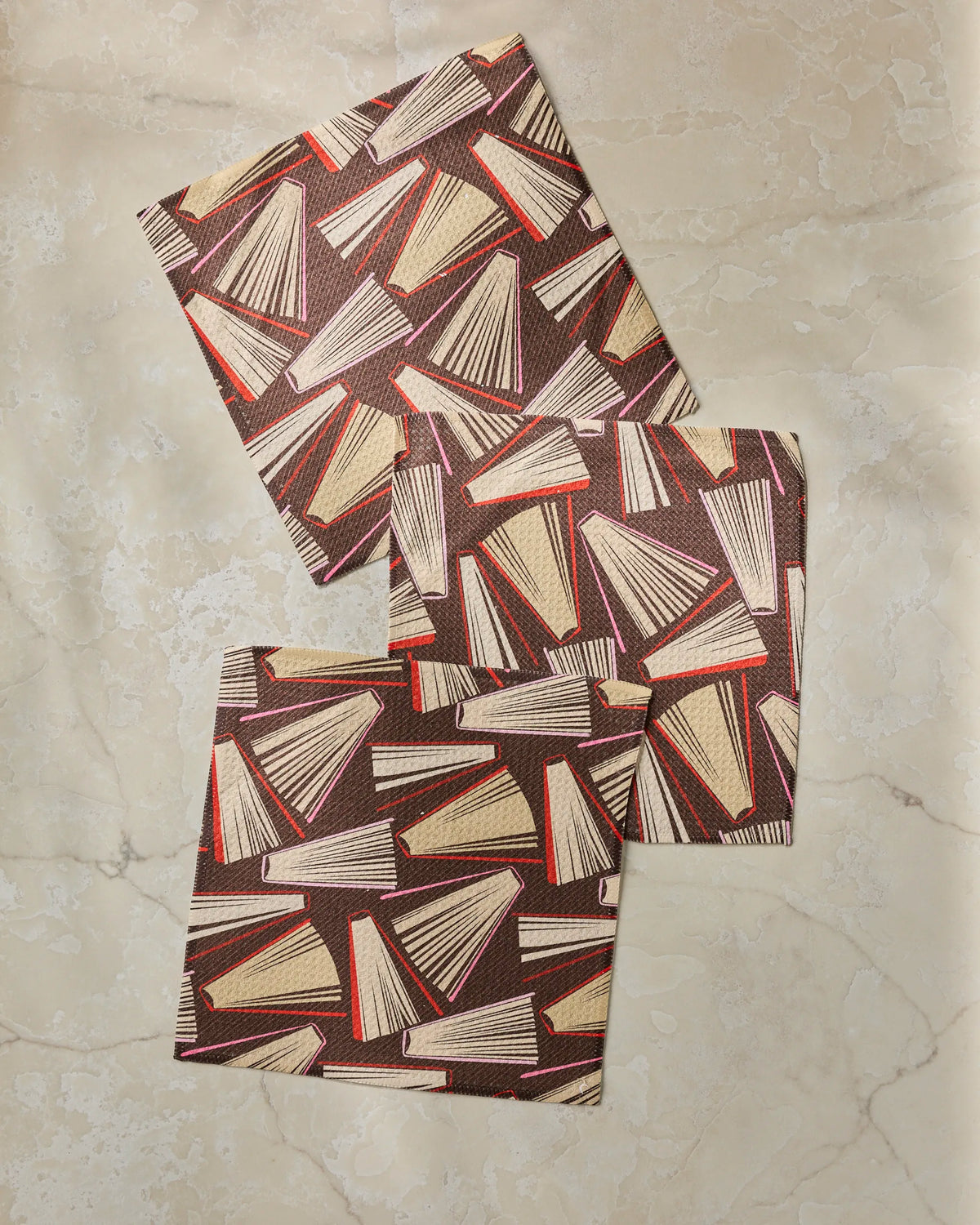 Three square Bookmas fabric coasters by Geometry, featuring a geometric pattern in brown, red, and cream hues and crafted from recycled materials, are arranged on a beige marble surface.