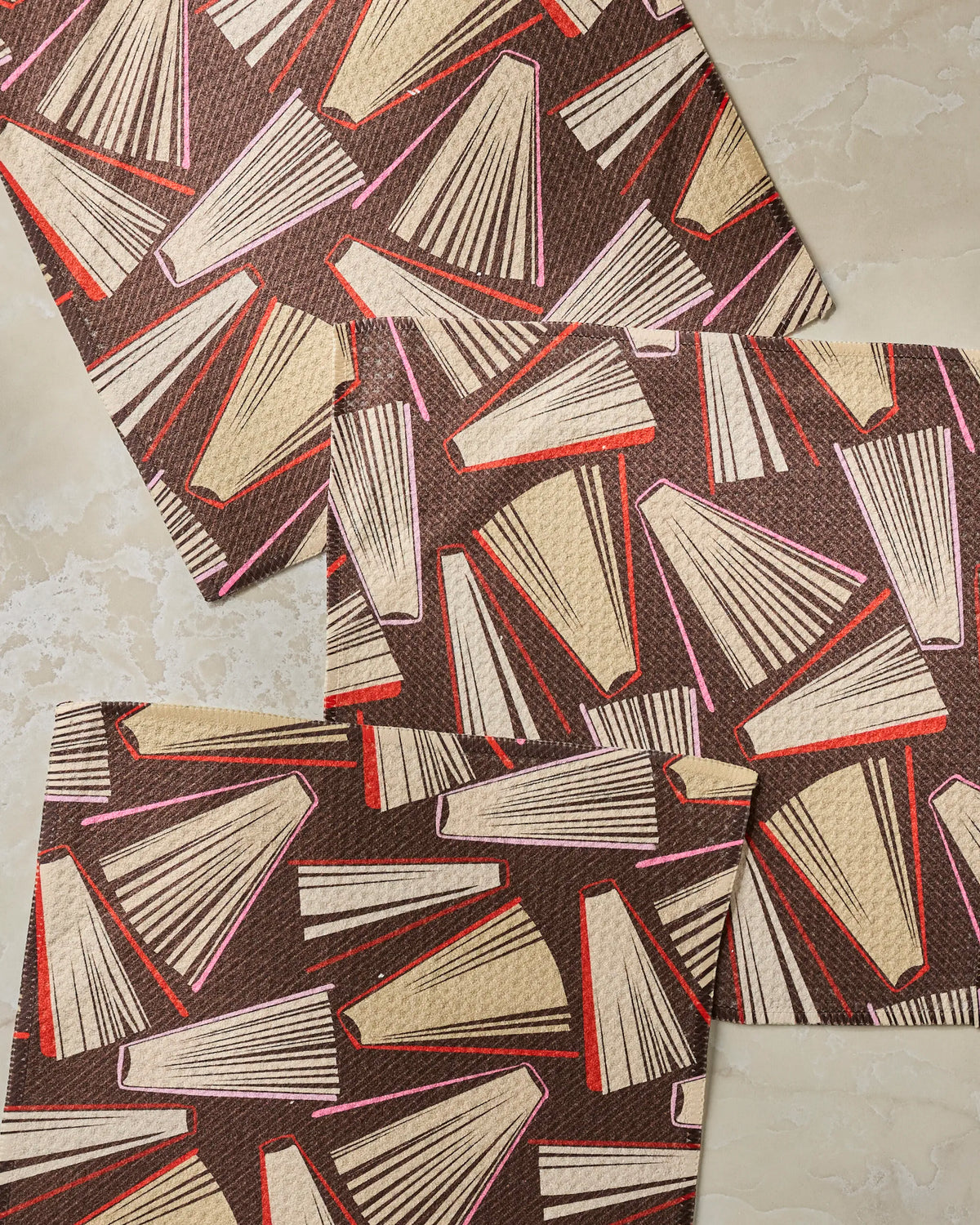 Three rectangular Bookmas mats by Geometry, made from recycled materials and featuring a repeating pattern of closed books in beige, red, and pink on a brown background, arranged on a light, textured surface.