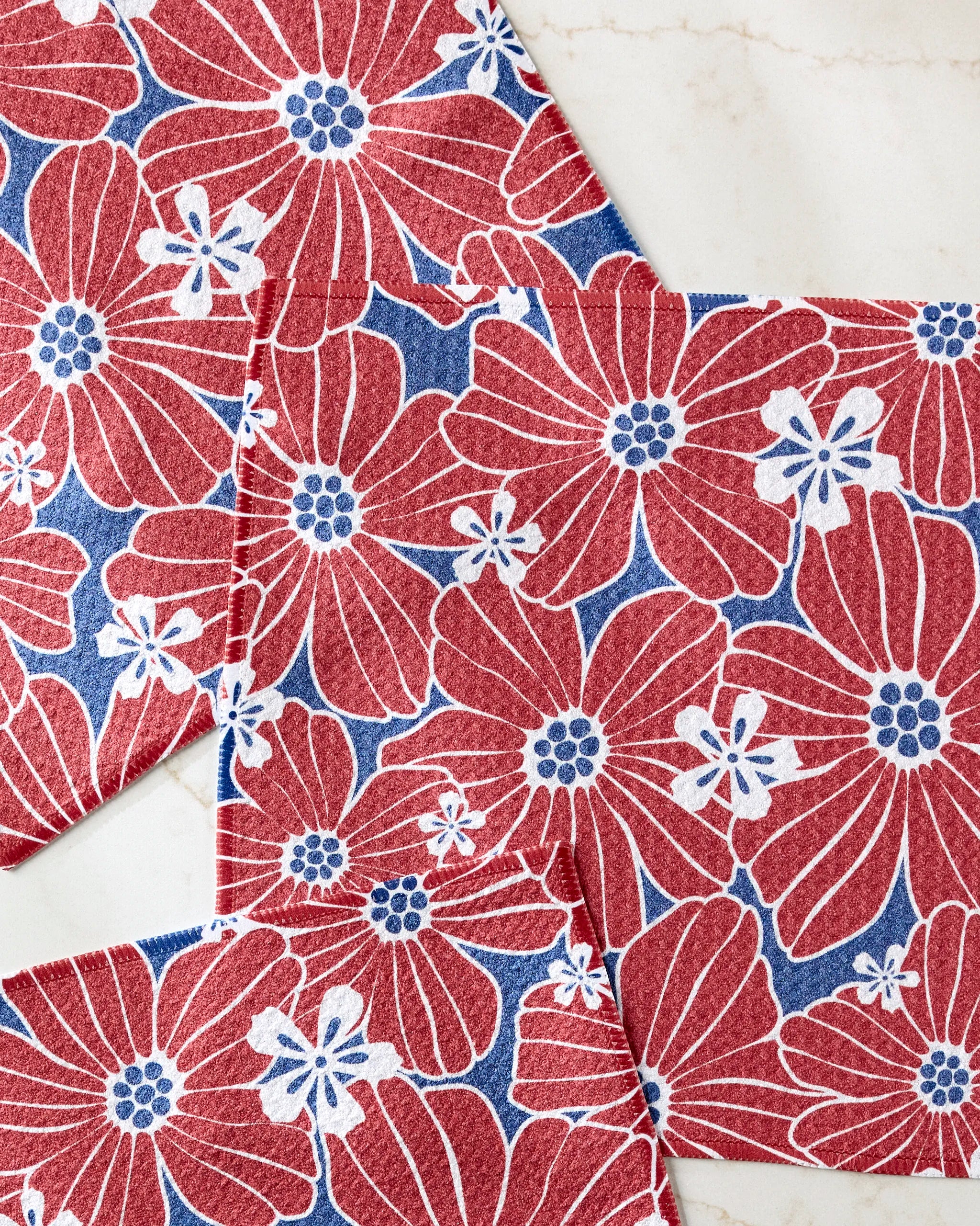 Botanical Blush napkins by Ashley Celena, with red and white florals on blue, overlap elegantly on marble—adding vibrant decor to your kitchen. Ideal for use in colorful dishcloth sets or as absorbent dishcloths.