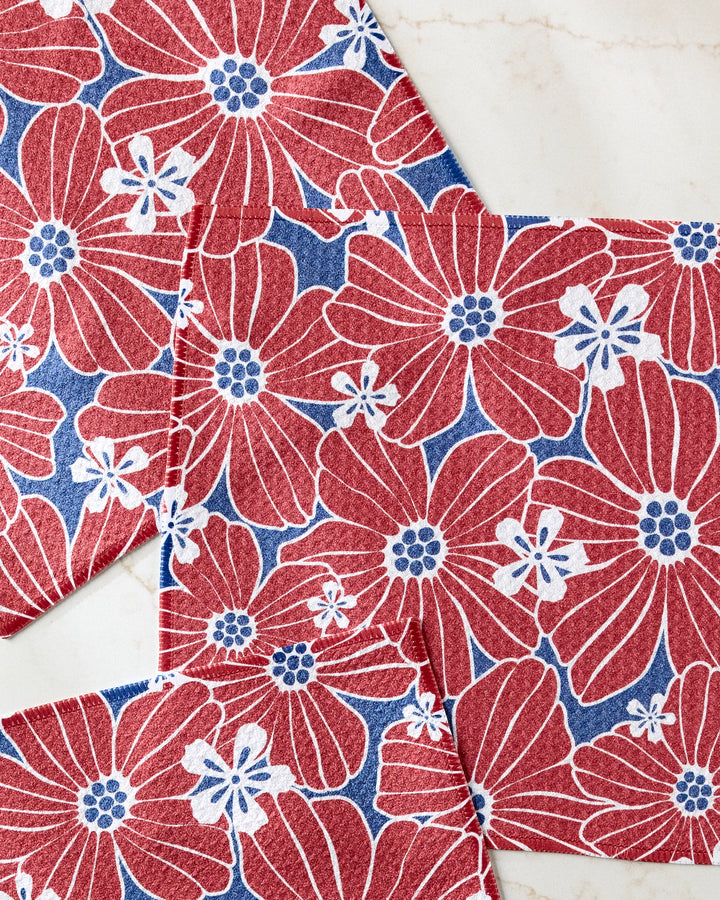 Botanical Blush napkins by Ashley Celena, with red and white florals on blue, overlap elegantly on marble—adding vibrant decor to your kitchen. Ideal for use in colorful dishcloth sets or as absorbent dishcloths.