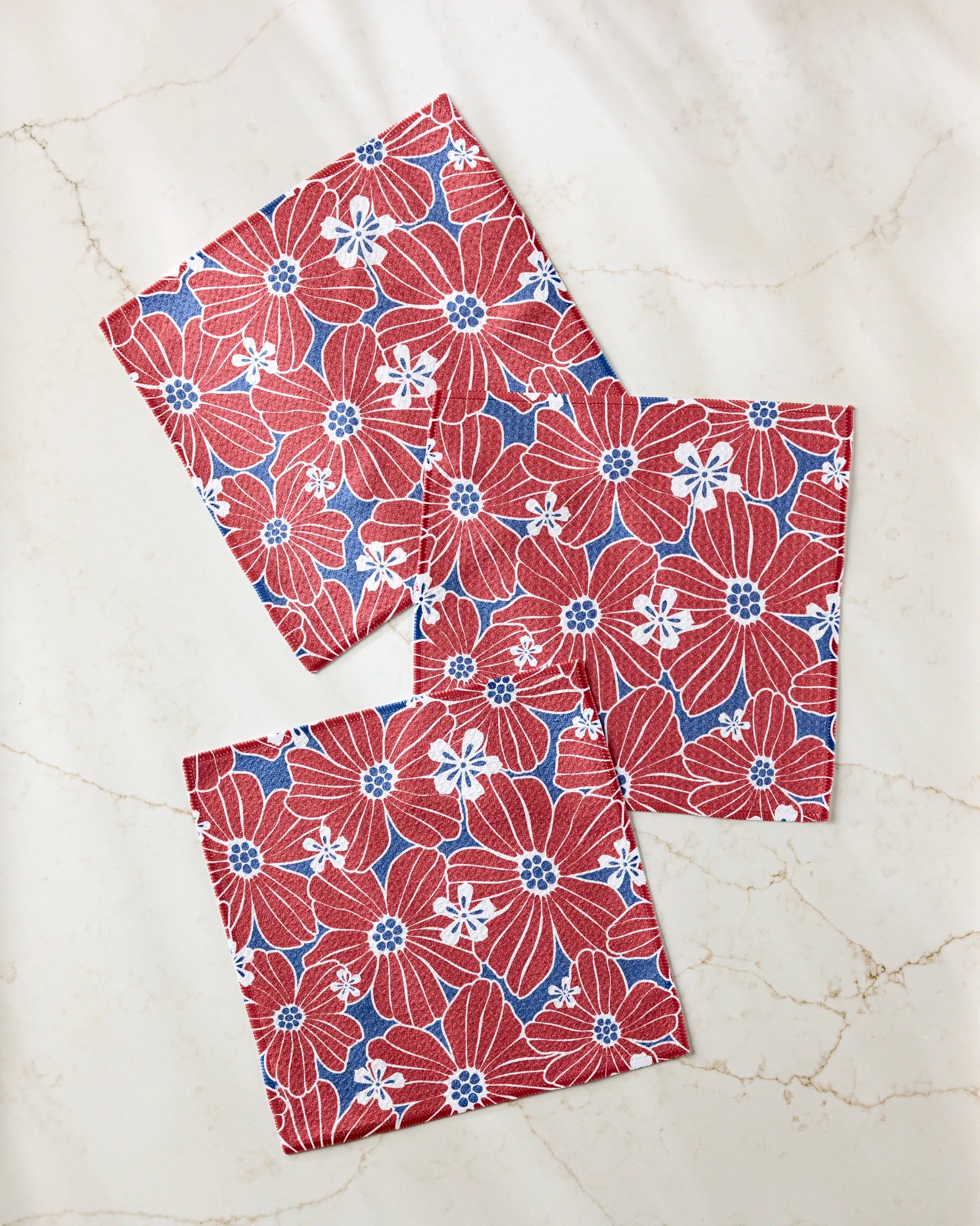Three square Botanical Blush dishcloths by Ashley Celena display a red background with a white and blue floral pattern, arranged diagonally and slightly overlapping on a light marbled surface.