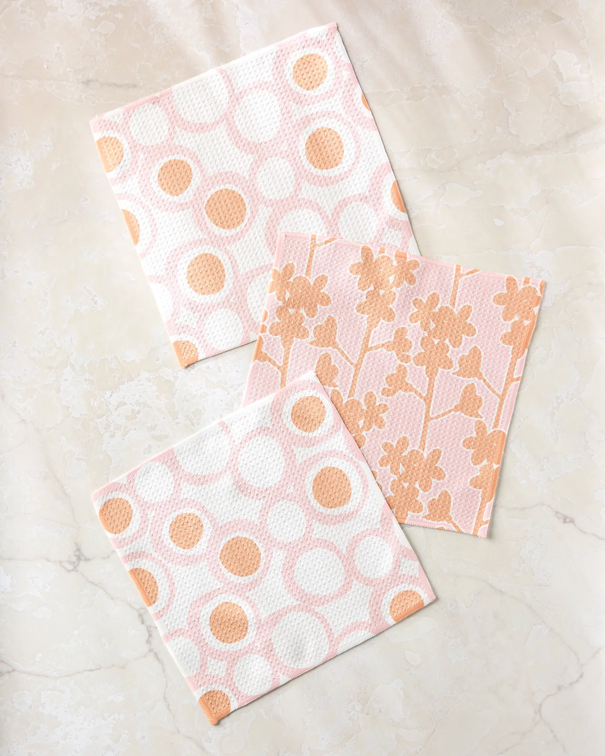Three Bouncing Balls cleaning cloths by Lizzie Clark, made from recycled materials, rest on a marble surface; two have polka dots and one features a floral design in peach and white.