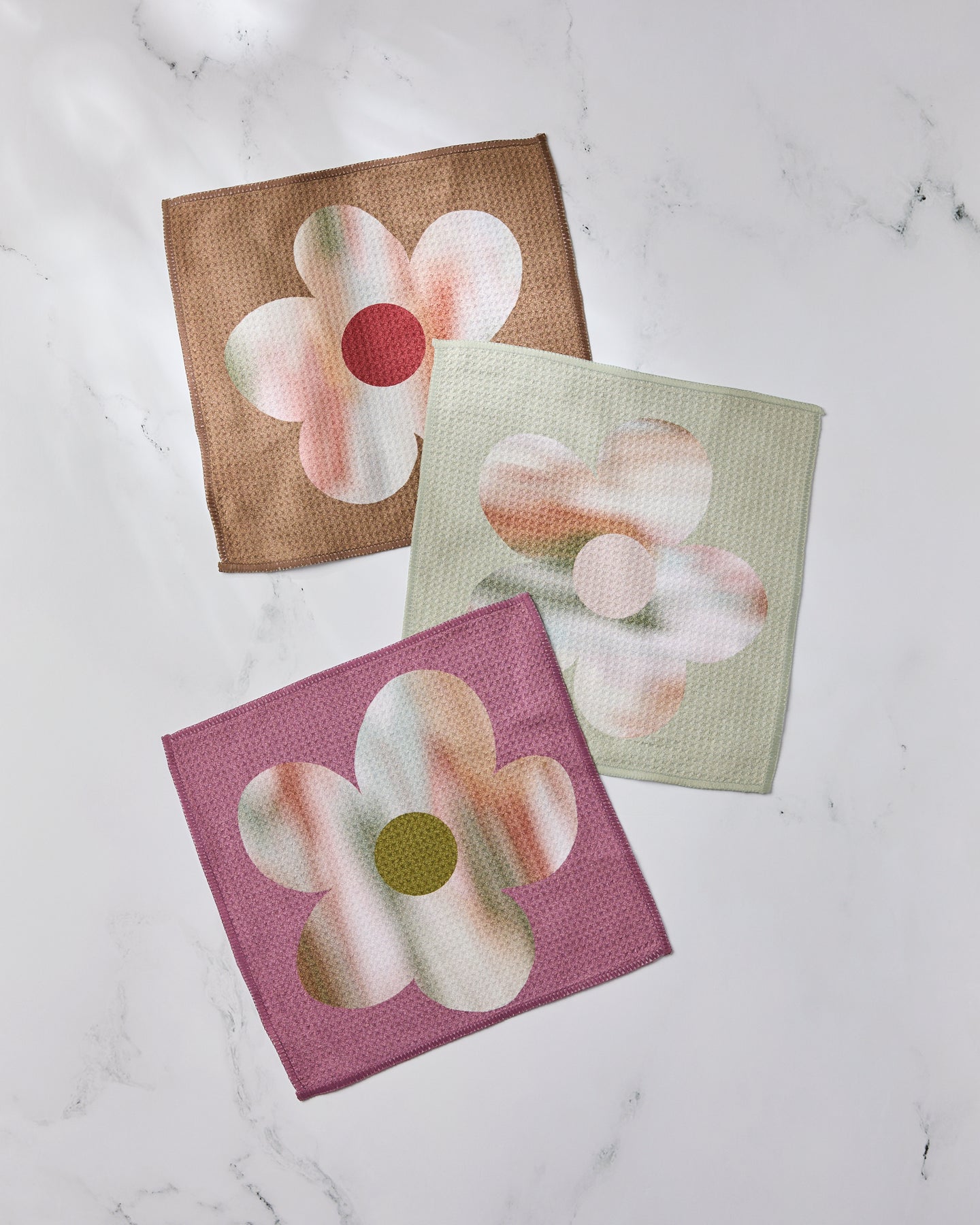 The Geometry Brushed Bouquet set features three textured, super-absorbent dishtowels with abstract flower designs in brown with red, green with orange, and purple with green accents on a white marble background.