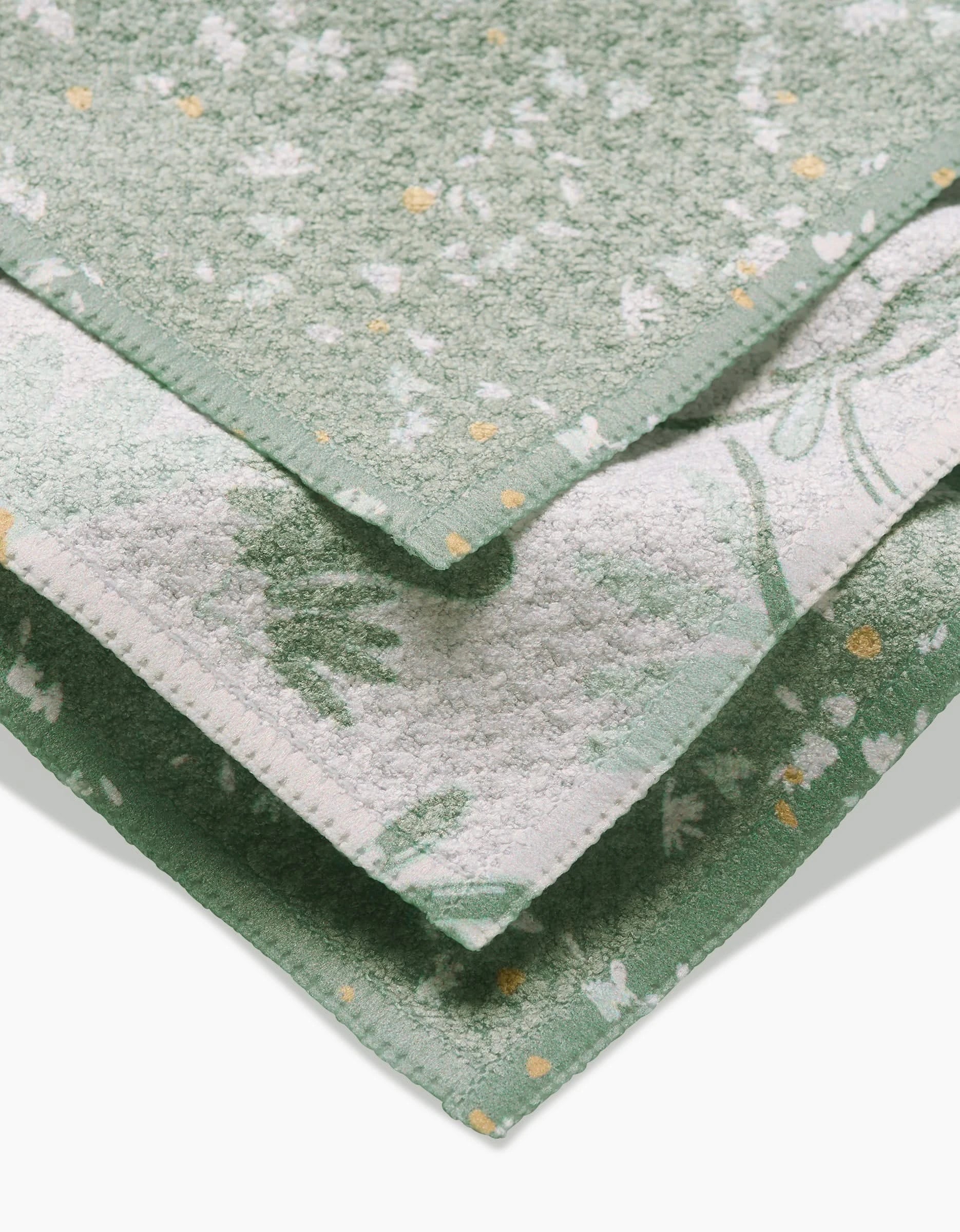 Three soft, green Bunny Tea Time towels from Second Nature Studio are stacked neatly. Made of absorbent Geoweave fabric, each towel features unique white and yellow floral patterns and different edge designs on each layer.