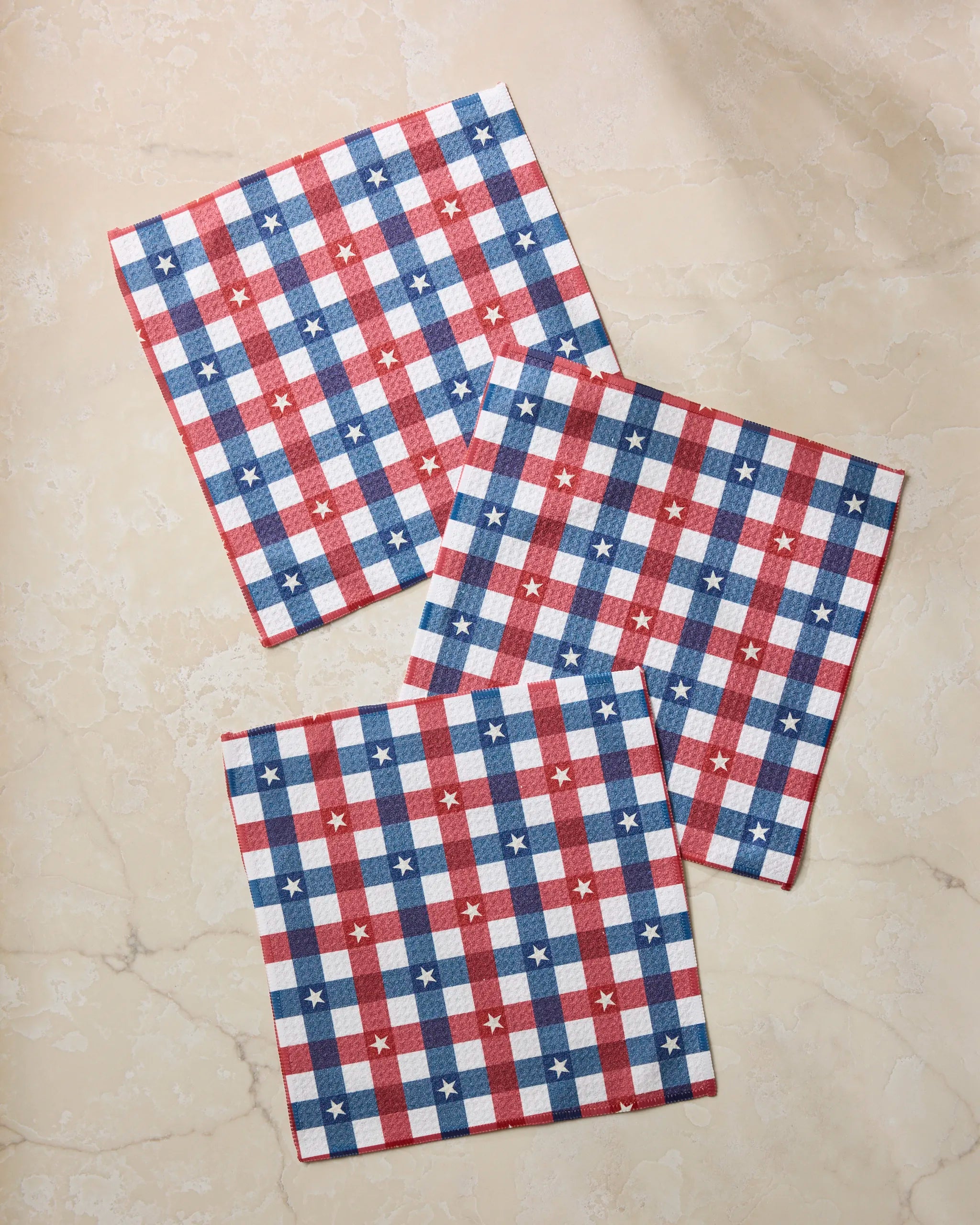 Three Geometry Bunting Check square napkins made from Geoweave fabric, with a red, white, and blue checkered pattern and small white stars, are displayed on a beige marble surface.