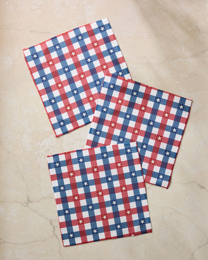Three Geometry Bunting Check square napkins made from Geoweave fabric, with a red, white, and blue checkered pattern and small white stars, are displayed on a beige marble surface.