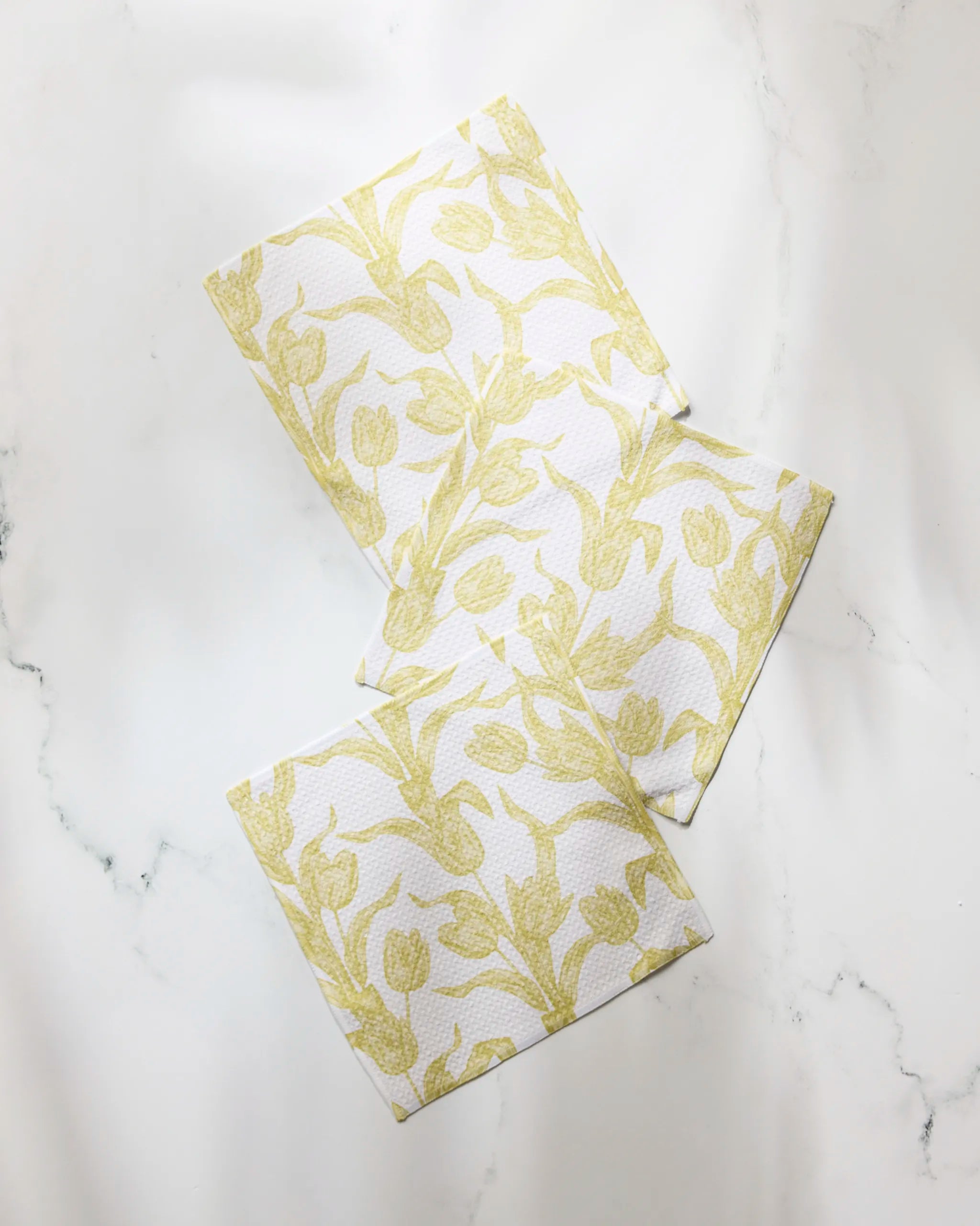 Three Geometry Butter Tulips dishcloths, featuring a yellow floral pattern and made from recycled materials, are arranged overlapping on a white marble surface.