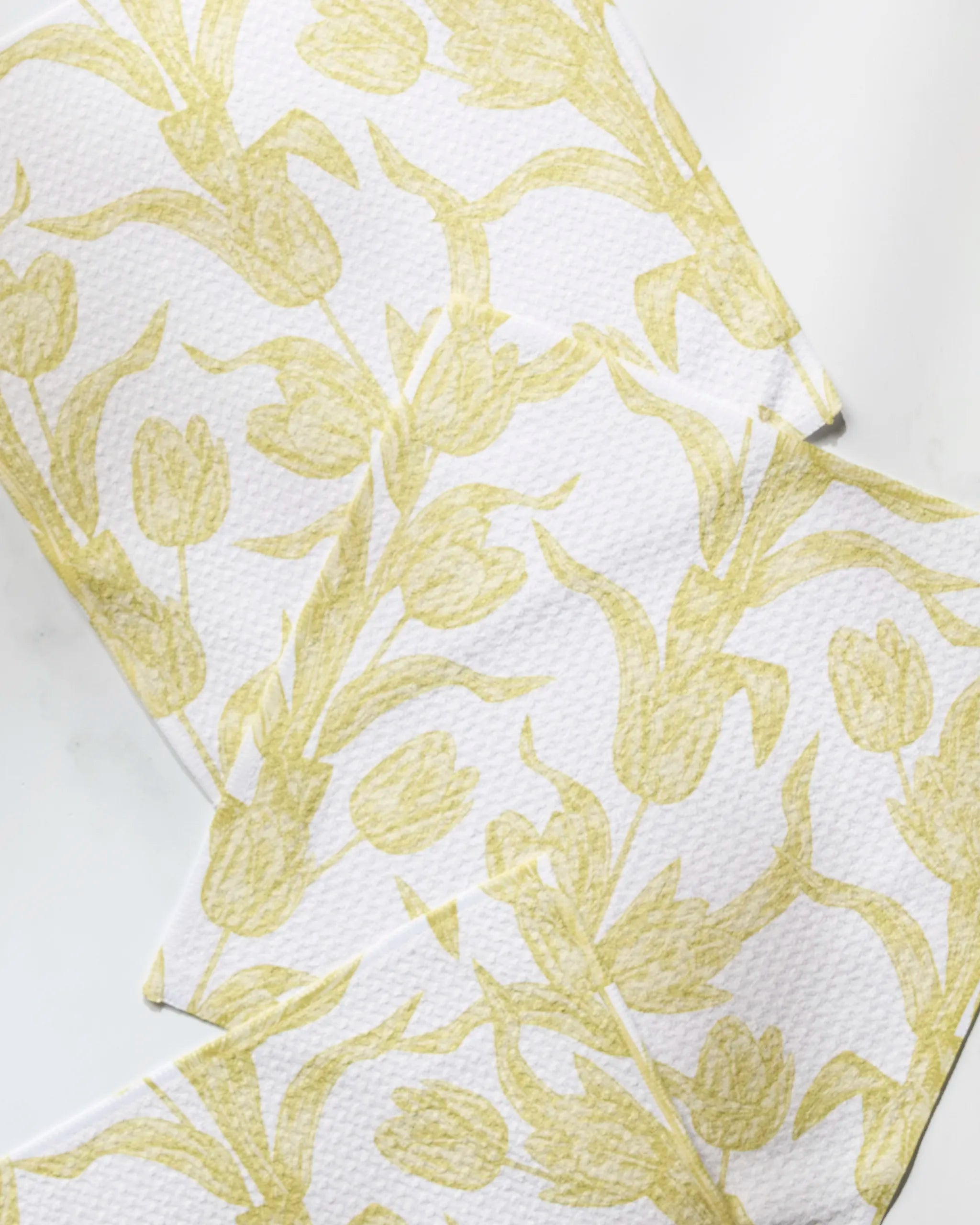 Three white cloths from Geometry’s Butter Tulips set, featuring a pale yellow tulip pattern, are arranged overlapping on a white surface—charming dishcloths made from recycled materials.