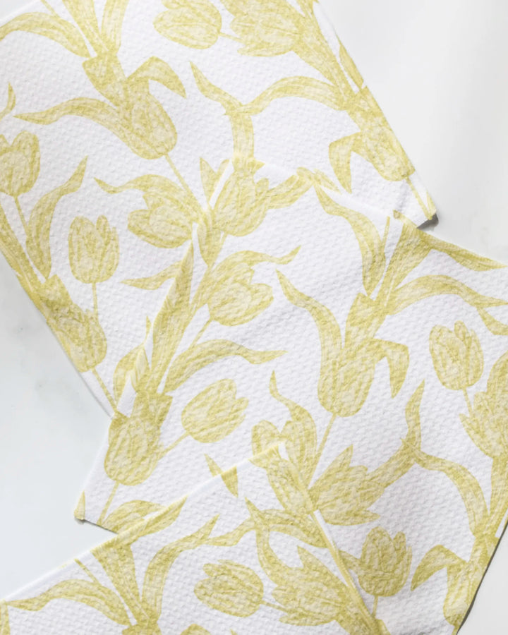 Three white cloths from Geometry’s Butter Tulips set, featuring a pale yellow tulip pattern, are arranged overlapping on a white surface—charming dishcloths made from recycled materials.