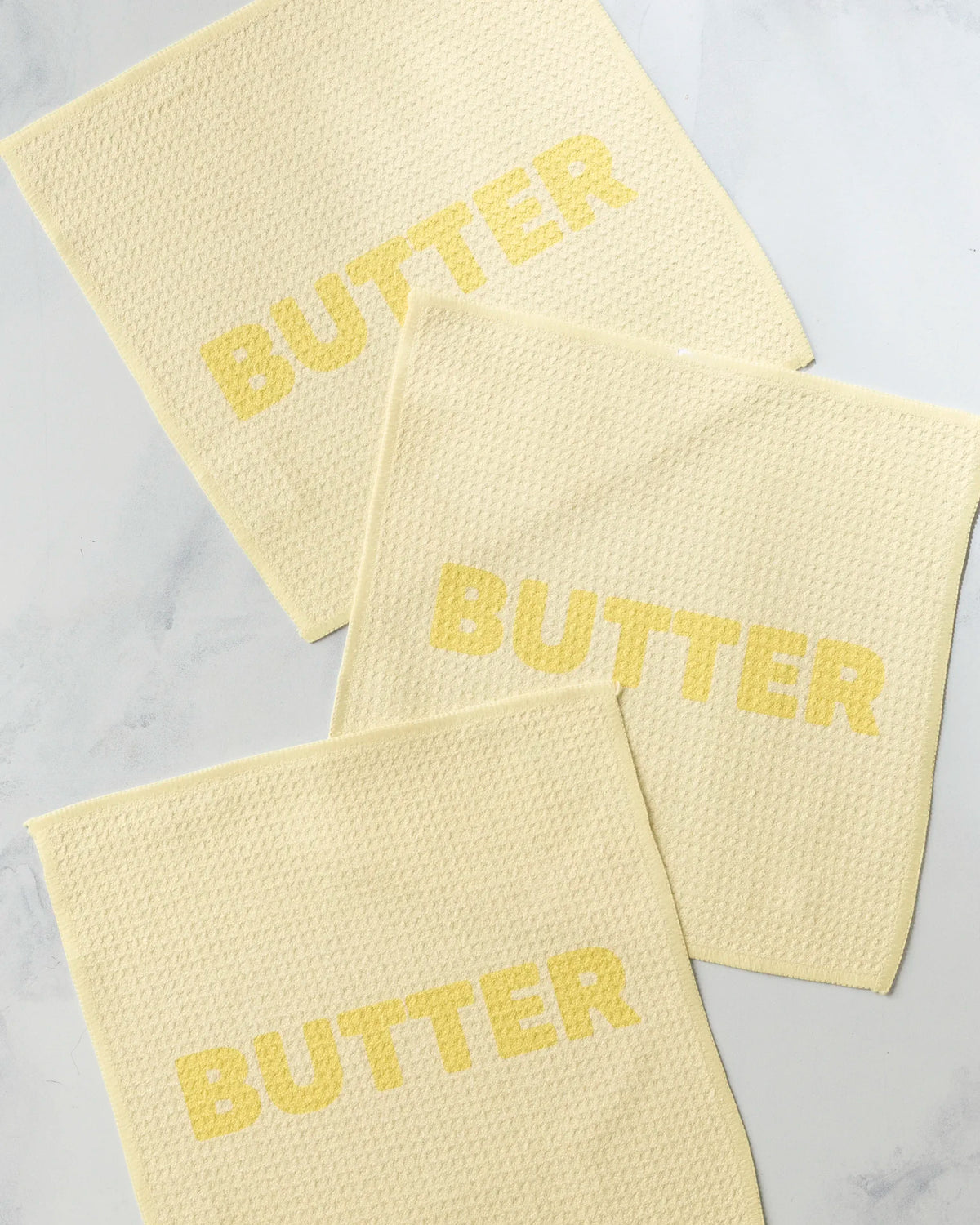 Three pale yellow, super-absorbent GEOMETRY Butter dishcloths with a textured square weave, each printed with the word BUTTER in a darker yellow font.