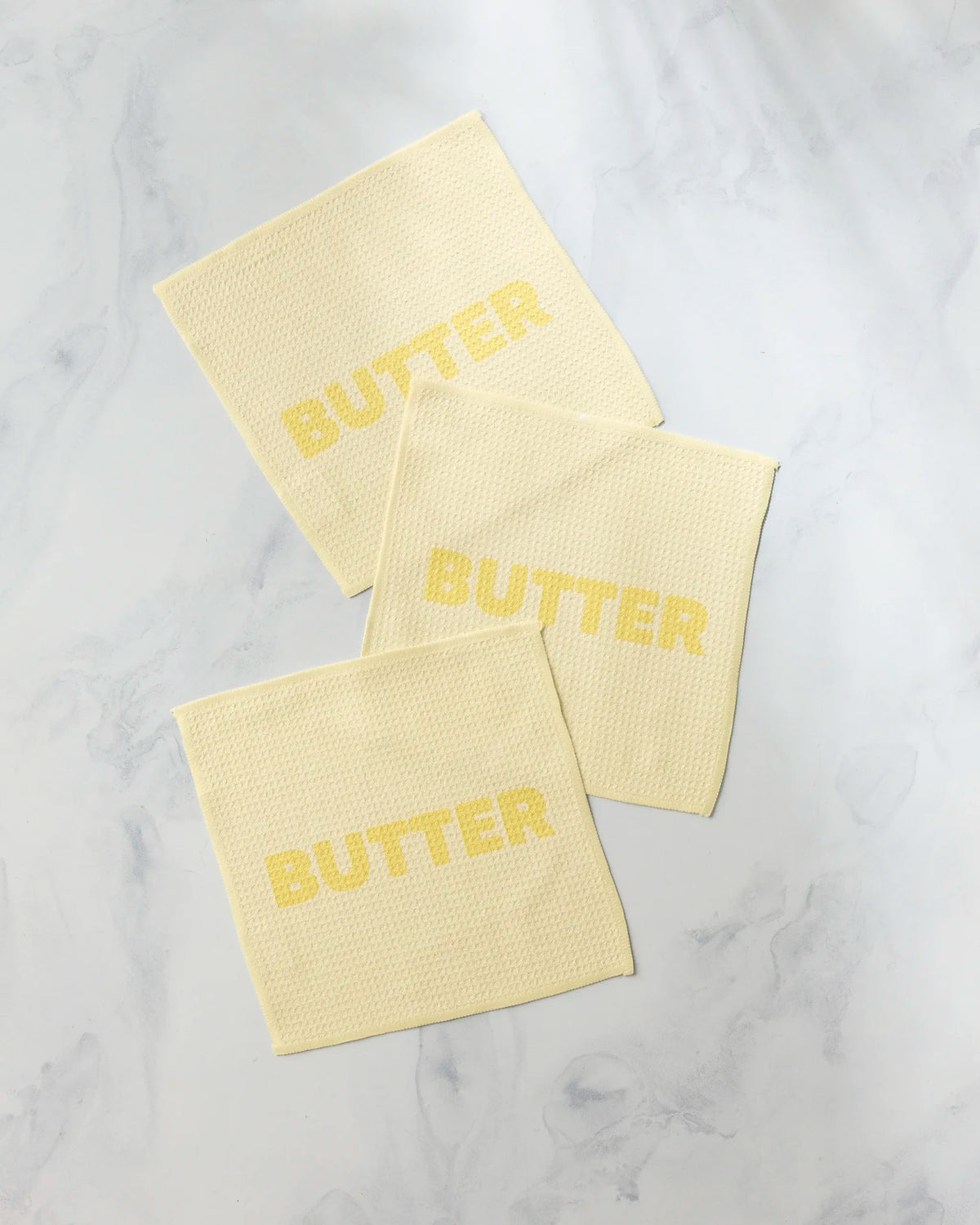 Three square, pale yellow, super-absorbent Geometry Butter dishcloths each featuring the word BUTTER printed in a darker yellow.