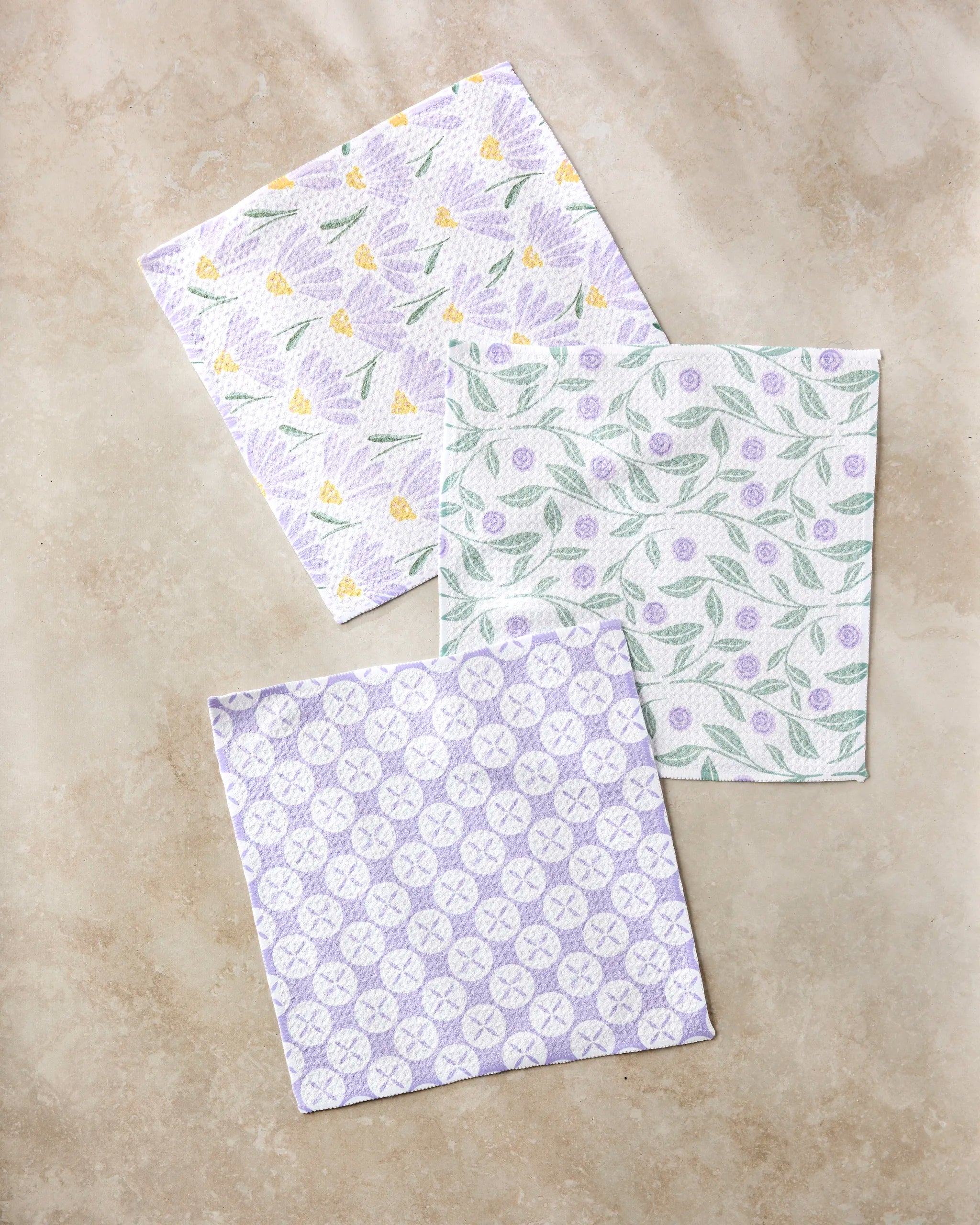 Button Vines and Daisies by Hufton Studio: three recycled square napkins with pastel florals—yellow and purple flowers, green leaves with purple buds, and a geometric purple pattern—on a light beige background.