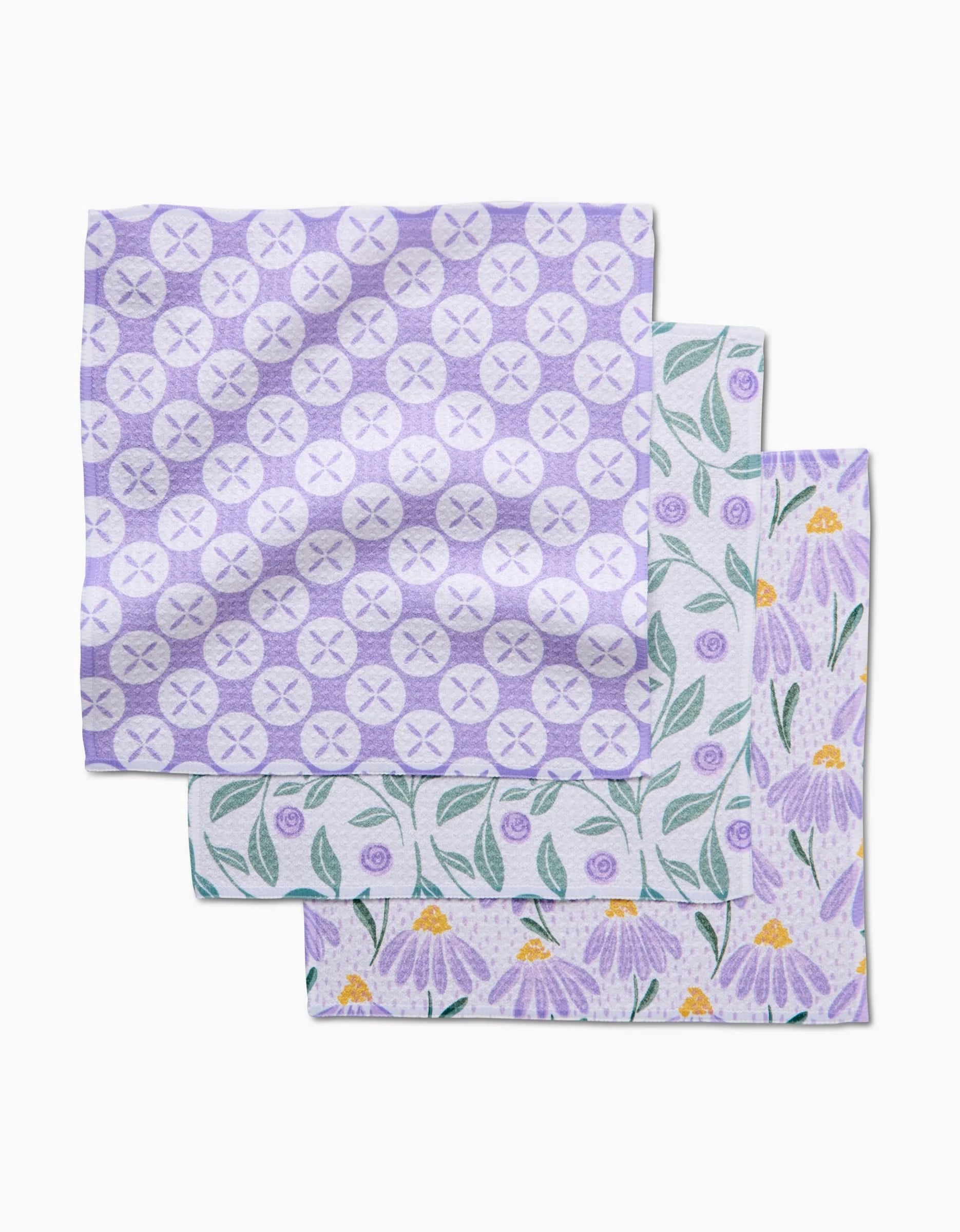 Three square Hufton Studio dishcloths from the Button Vines and Daisies set are stacked: the top is light purple with circles and Xs, while the others have purple flowers and green leaves on white Geoweave fabric.