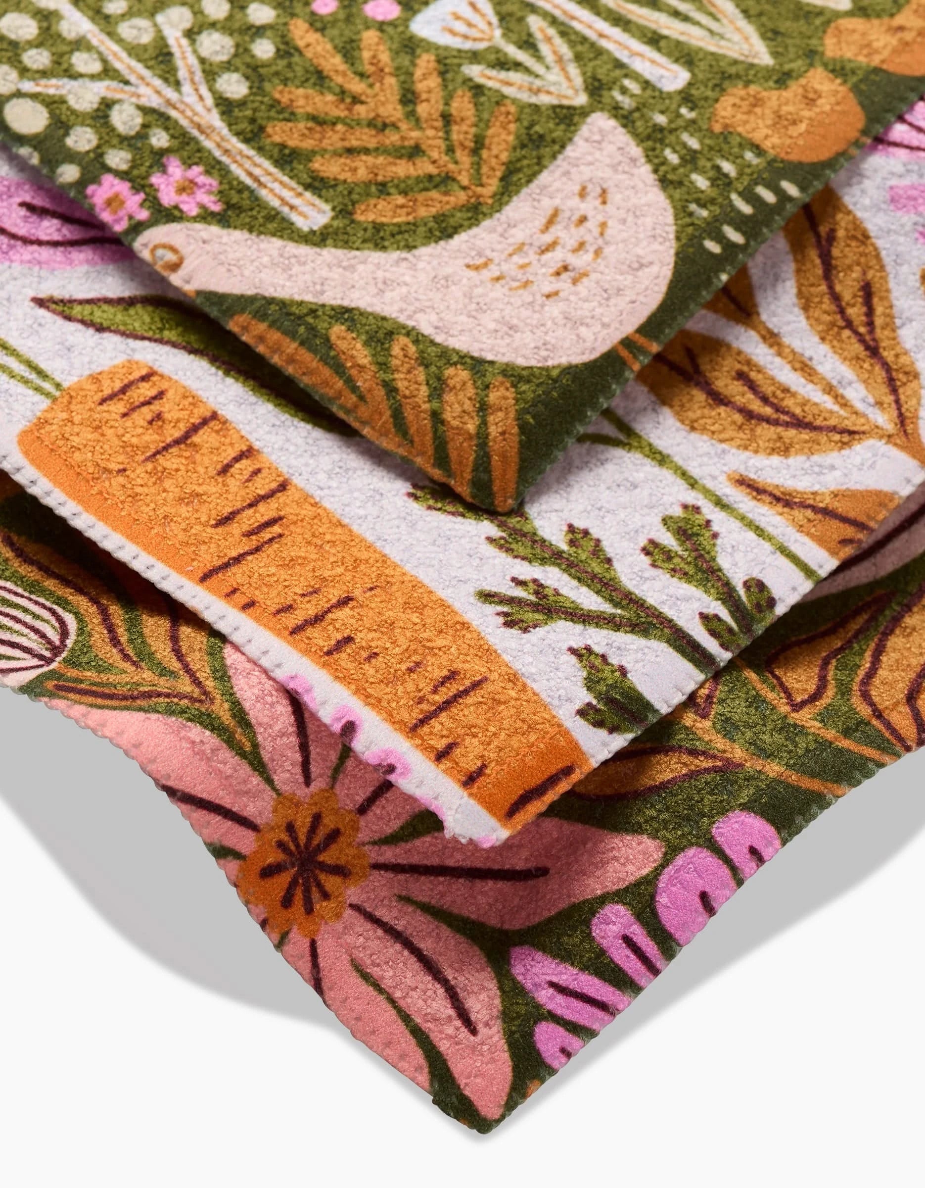 Close-up of three stacked Callagreen dishcloths by Amy MacCready, made from recycled materials with vibrant floral, leaf, and bird designs on green, orange, and pink Geoweave fabric backgrounds.