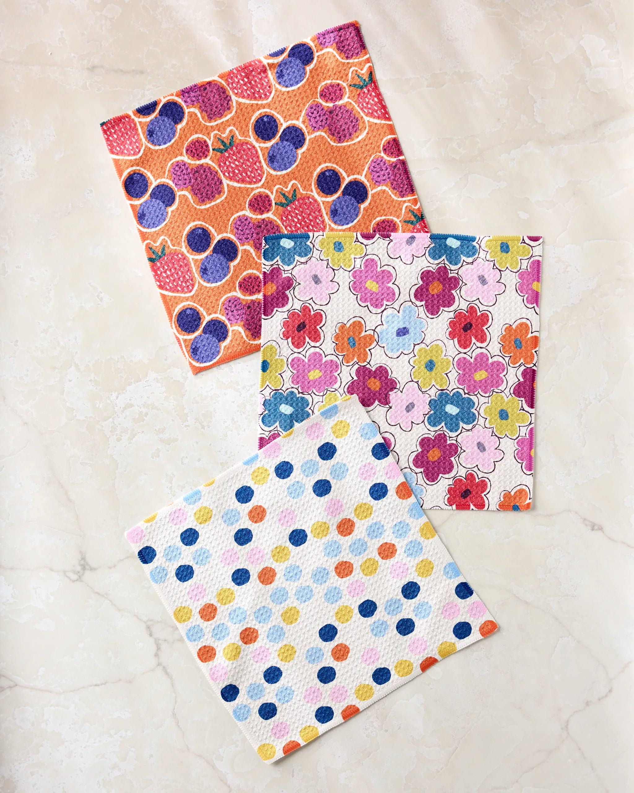 Three vibrant cloths from the Lizzie Clark Carnival Fling set are displayed on marble—one with strawberries, one with multicolored flowers, and one with blue, orange, and yellow dots.