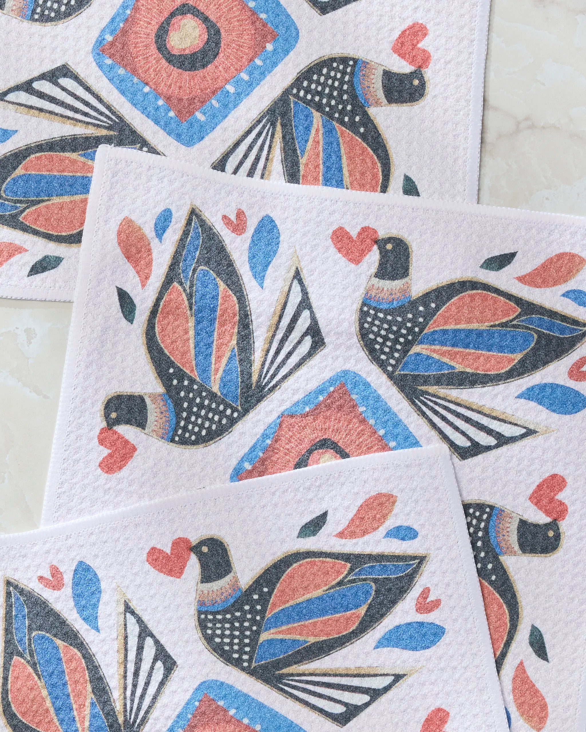Three white Geometry “Carrier” dishcloths, each featuring a colorful stylized bird in blue, red, or black with a red heart in its beak, are overlapped on a light background.