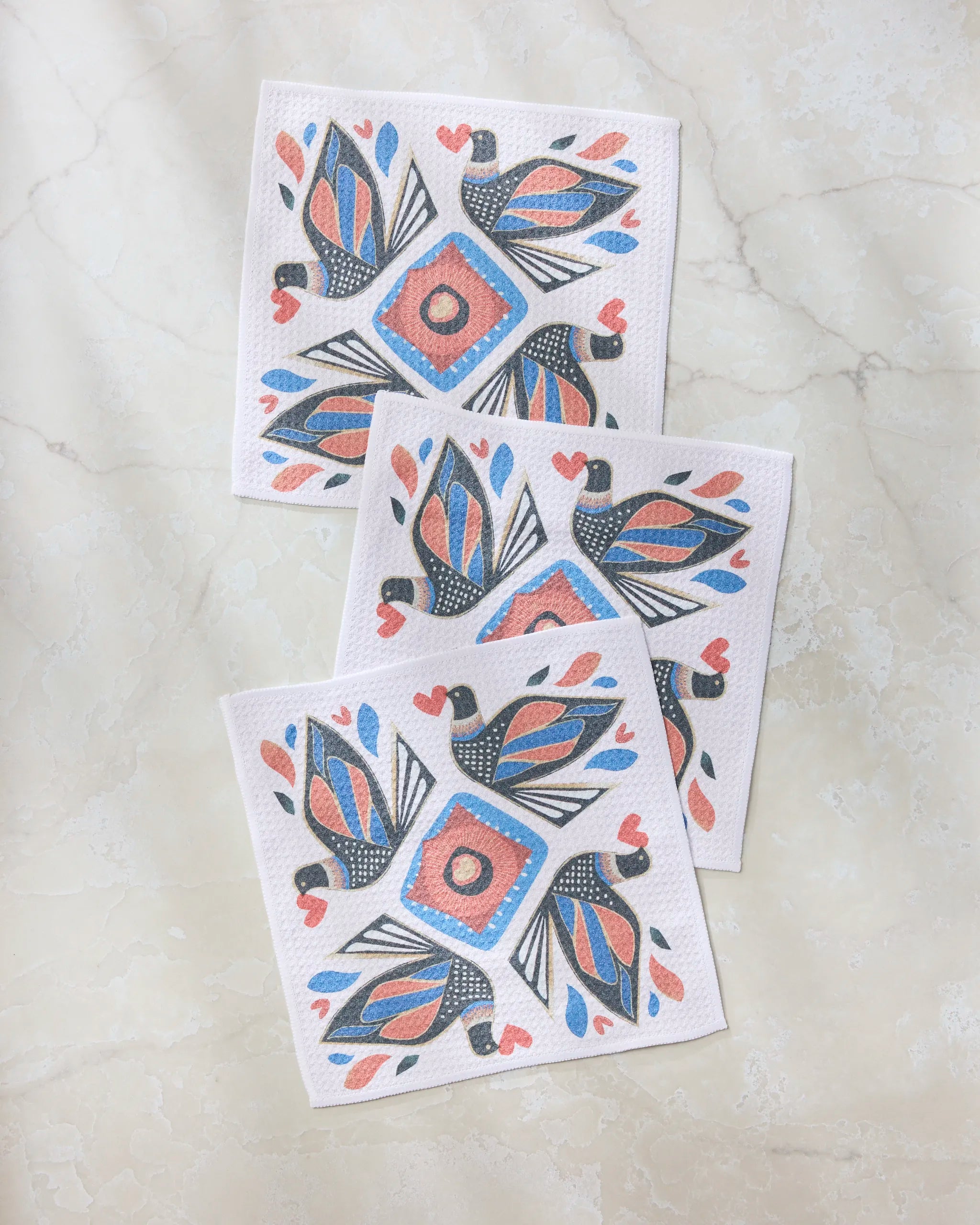 Three Geometry Carrier paper napkins featuring vibrant birds and geometric patterns in red, blue, and black are displayed on a light marble surface—perfect for pairing with your favorite super-absorbent dishcloths.