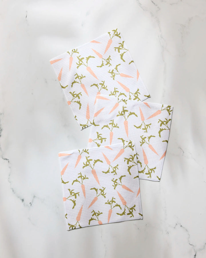 The Geometry Carrot Toss dishcloth set features three white napkins with a light pink carrot and green stem pattern, arranged on white marble—crafted from recycled materials for a charming, stylish, and eco-friendly touch.