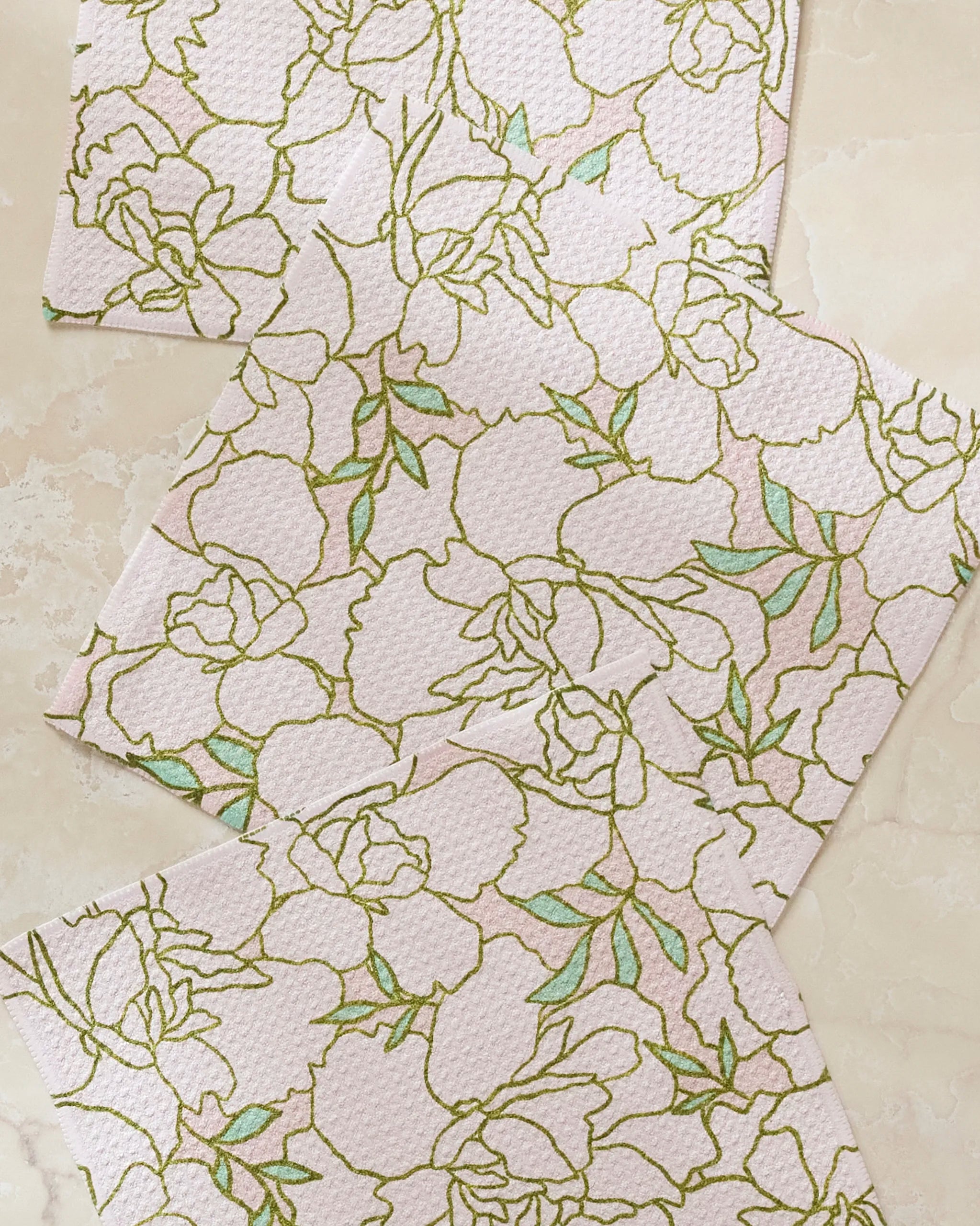 Three Celecia napkins by Amy MacCready, made from recycled materials, feature a pale pink textured surface and a delicate green and gold floral line pattern with large flowers and leaves. They are arranged on a light beige surface.