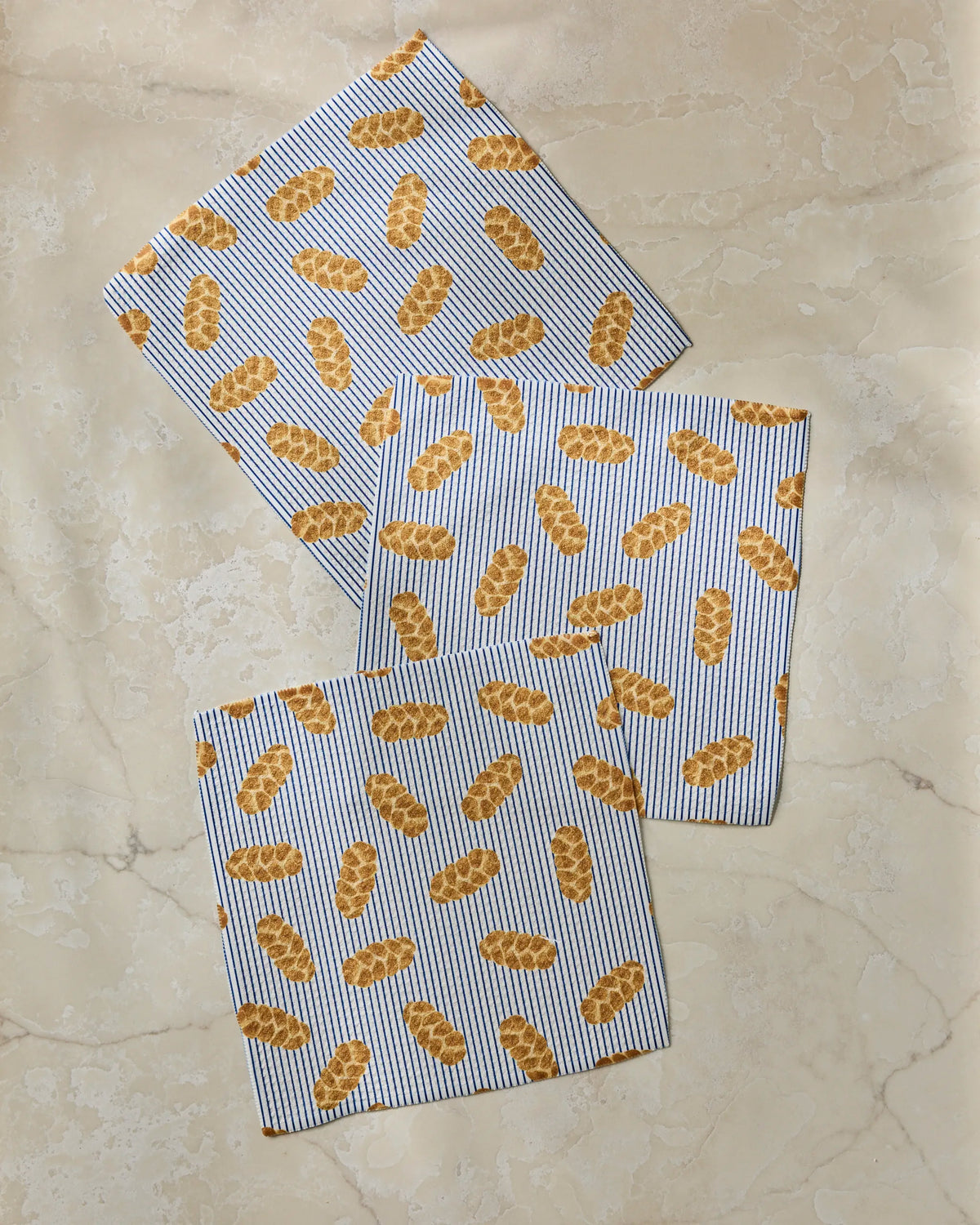 Three white “Challah Days” napkins from Geometry, featuring blue stripes and yellow bread loaf illustrations, are laid out on a beige marble surface.