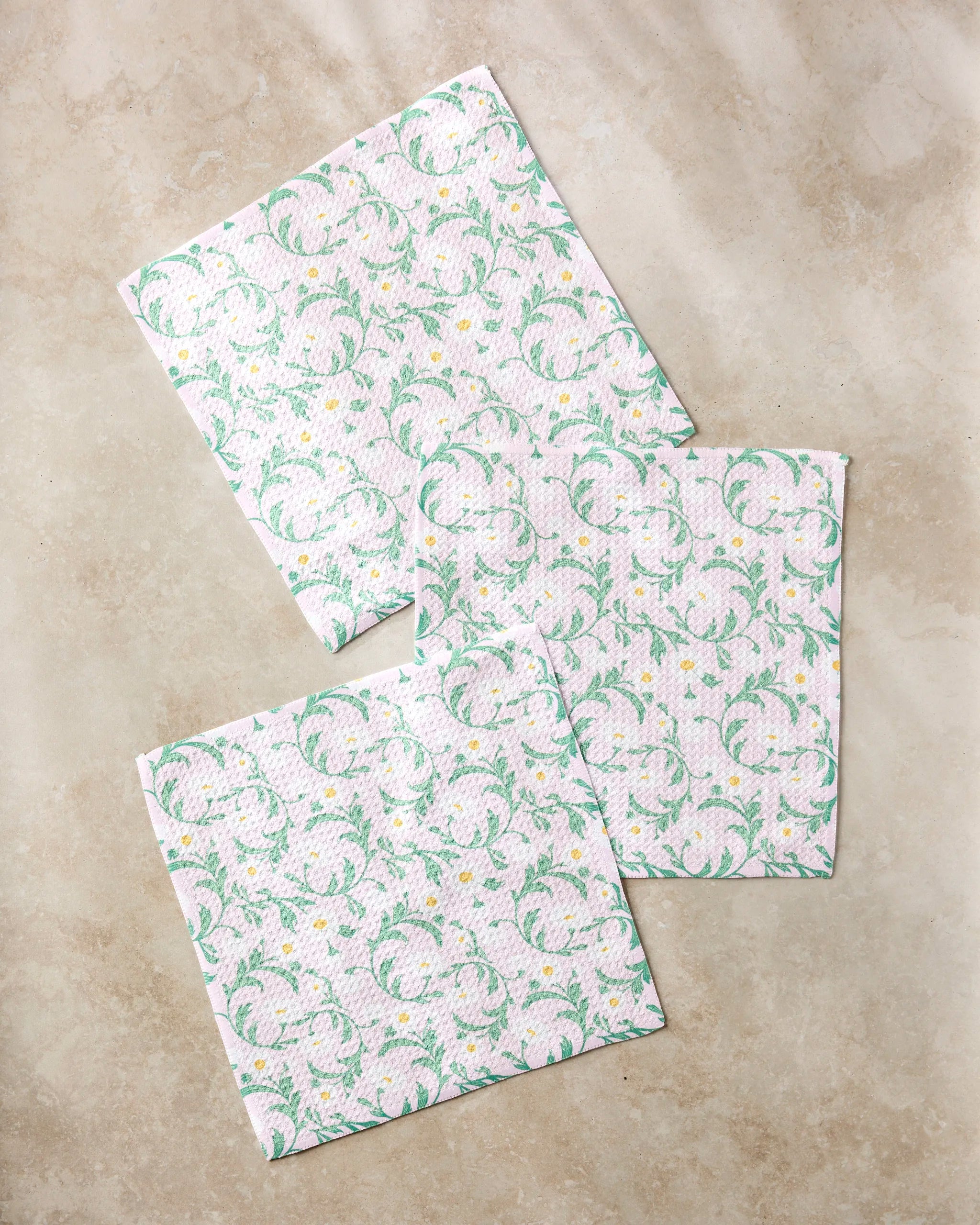 Three white super-absorbent Chamomille dishtowels by Danelys Sidron, featuring a green and yellow floral pattern, are arranged on a light beige textured surface.