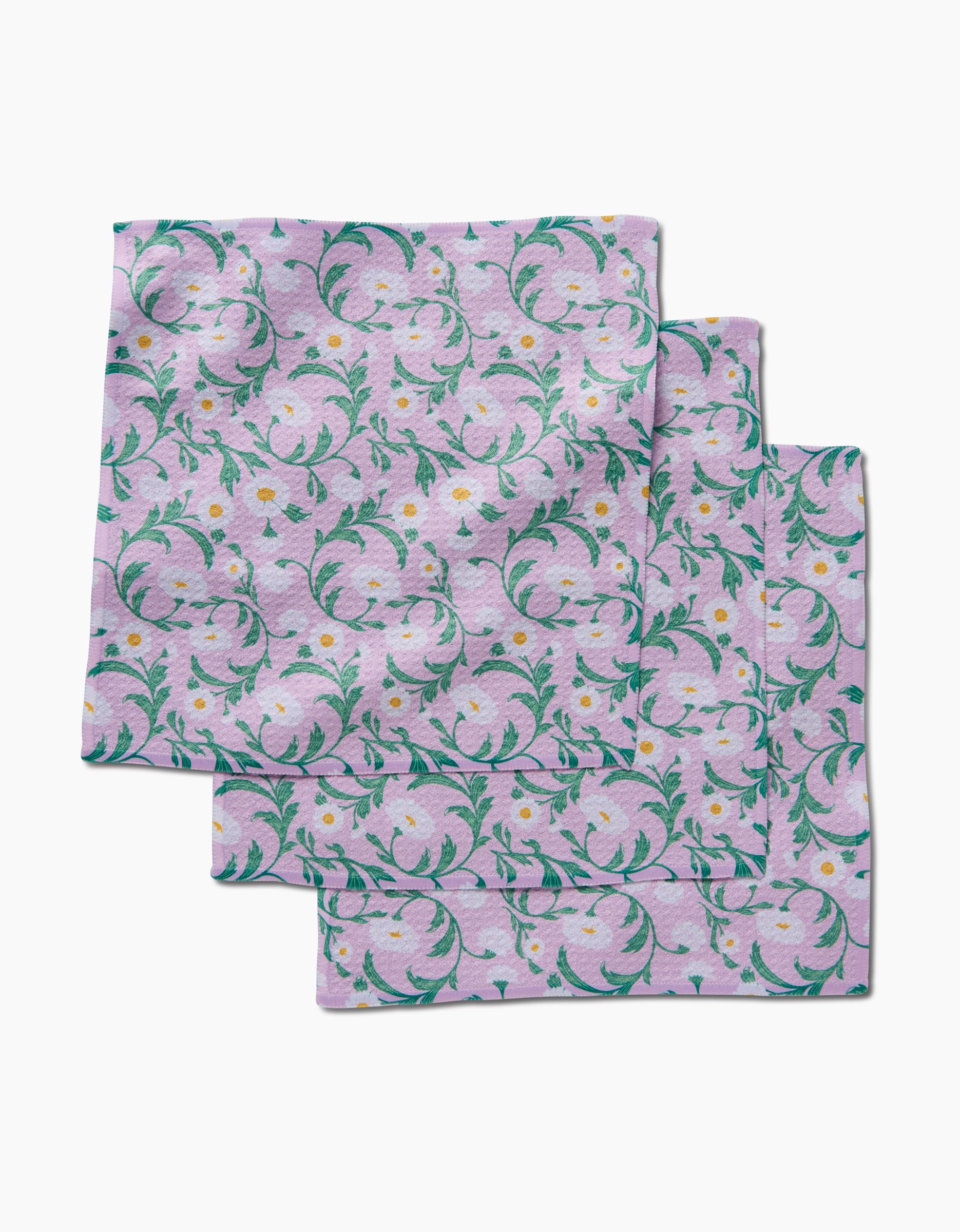 Three folded Chamomille dishcloths by Danelys Sidron, made from recycled materials, with a pink background and a repeating floral pattern of green leaves and small yellow flowers.