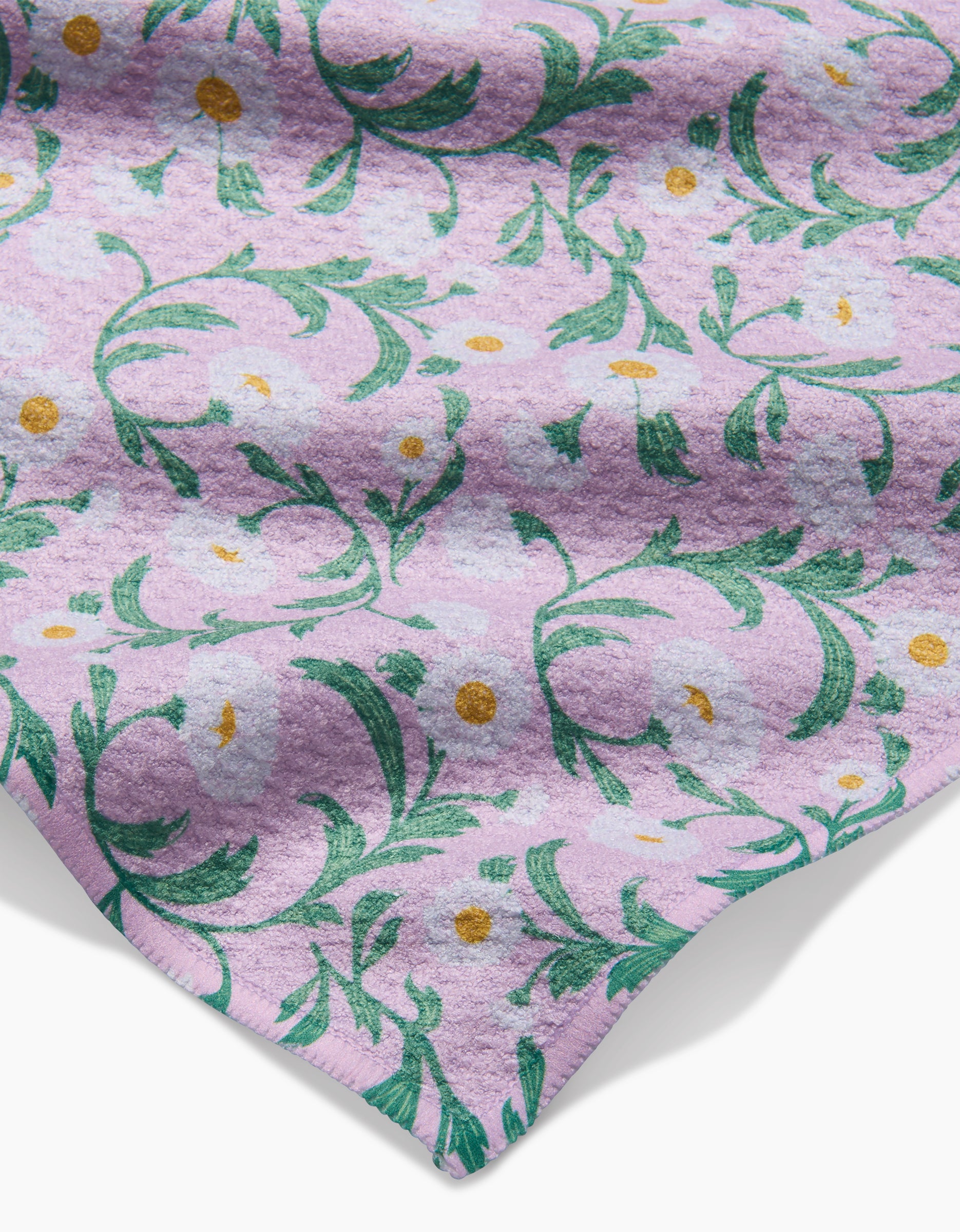 A close-up of Danelys Sidrons Chamomille dishcloth shows super-absorbent fabric with a light purple background, green floral pattern, white flowers, and small yellow dots. One corner of the cloth is slightly folded.