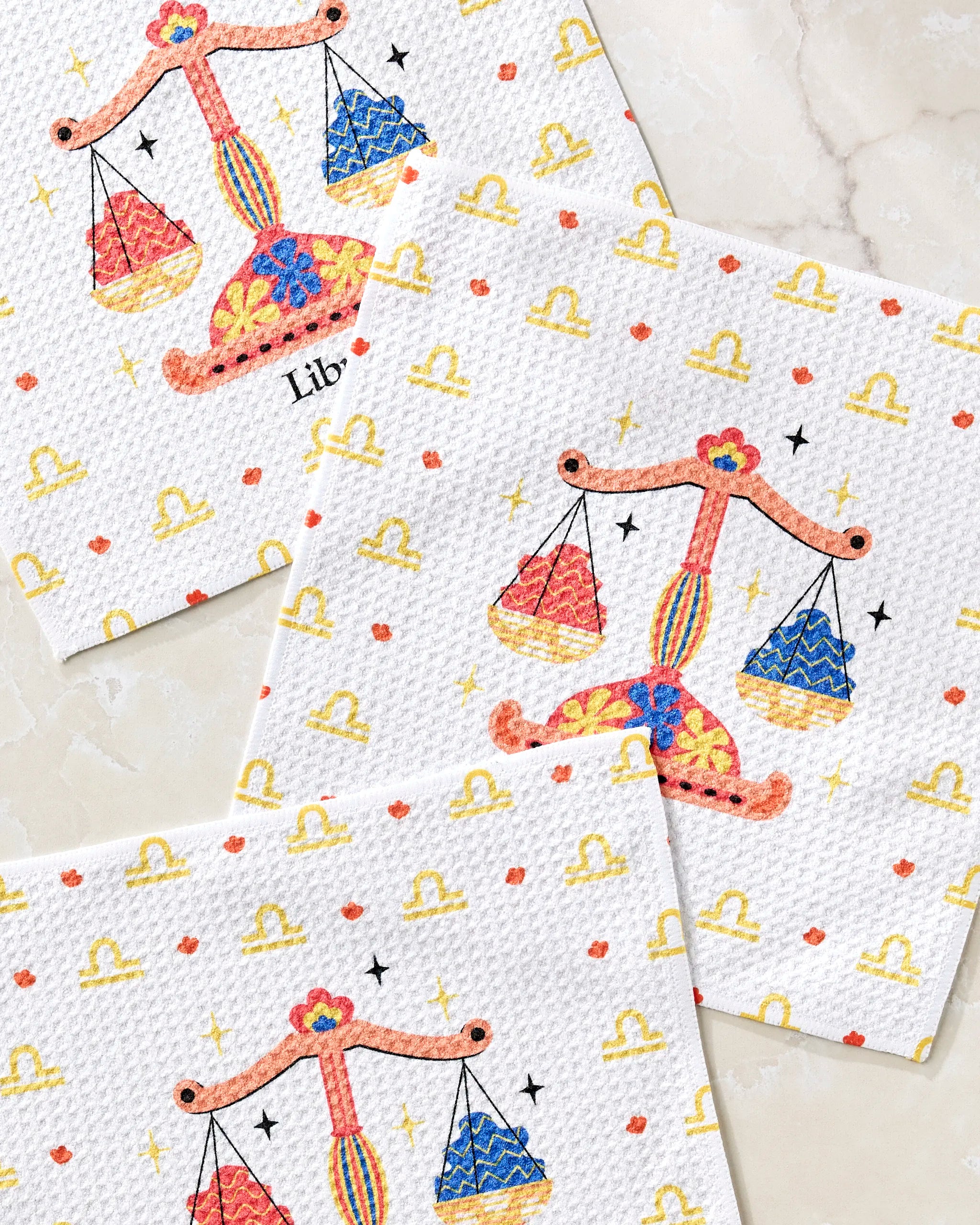 Three "Charming Libra" napkins by Heather Zhou, featuring colorful Libra zodiac scales, stars, and yellow symbols, are displayed on marble—perfect for pairing with your favorite dishcloth set.