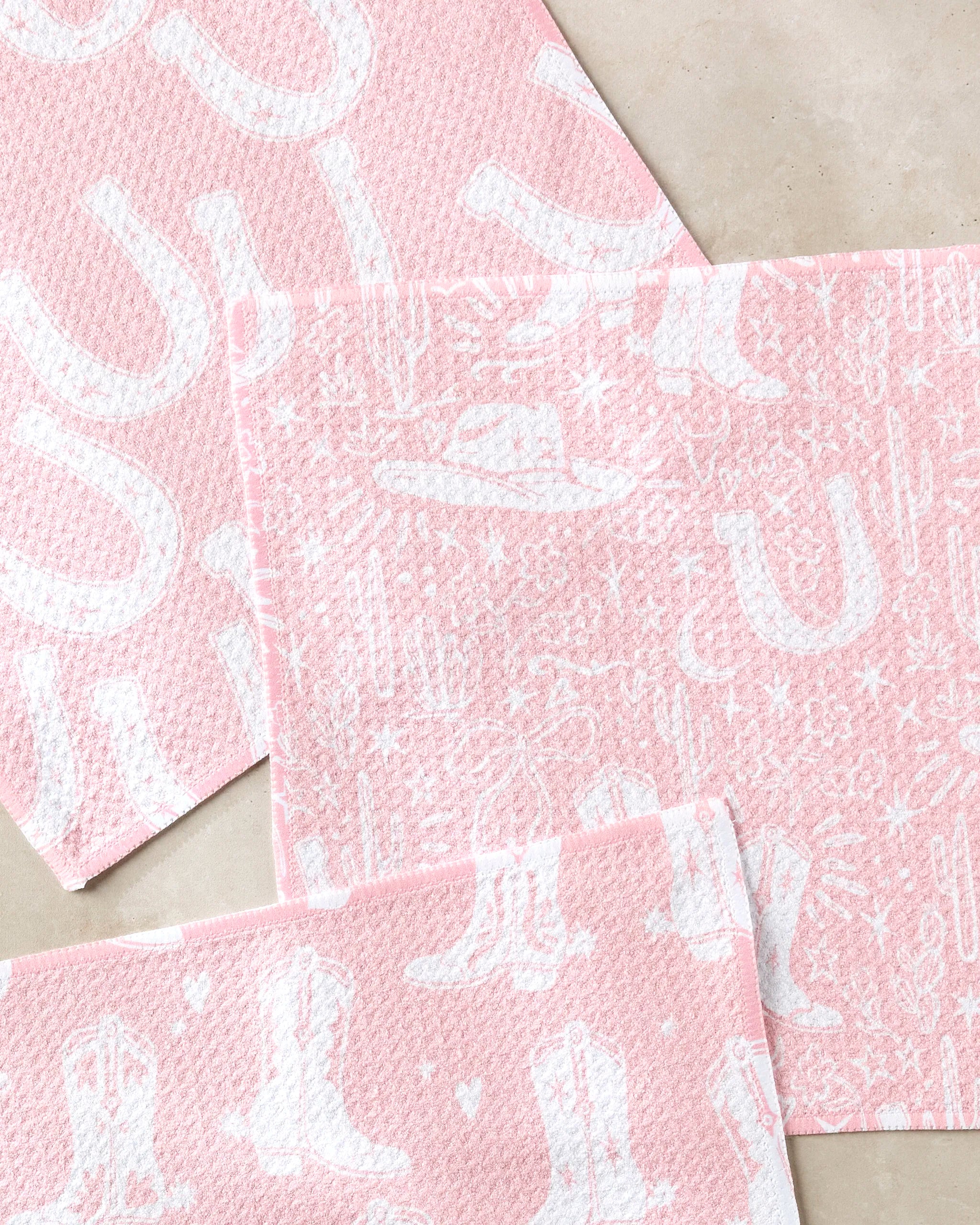 Three pink textured Chic Lucky Boots dishcloths by Aubrey Fairchild, featuring white Western-themed designs like cowboy boots, hats, cacti, stars, and horseshoes, are arranged on a beige surface for a fun and practical kitchen set.