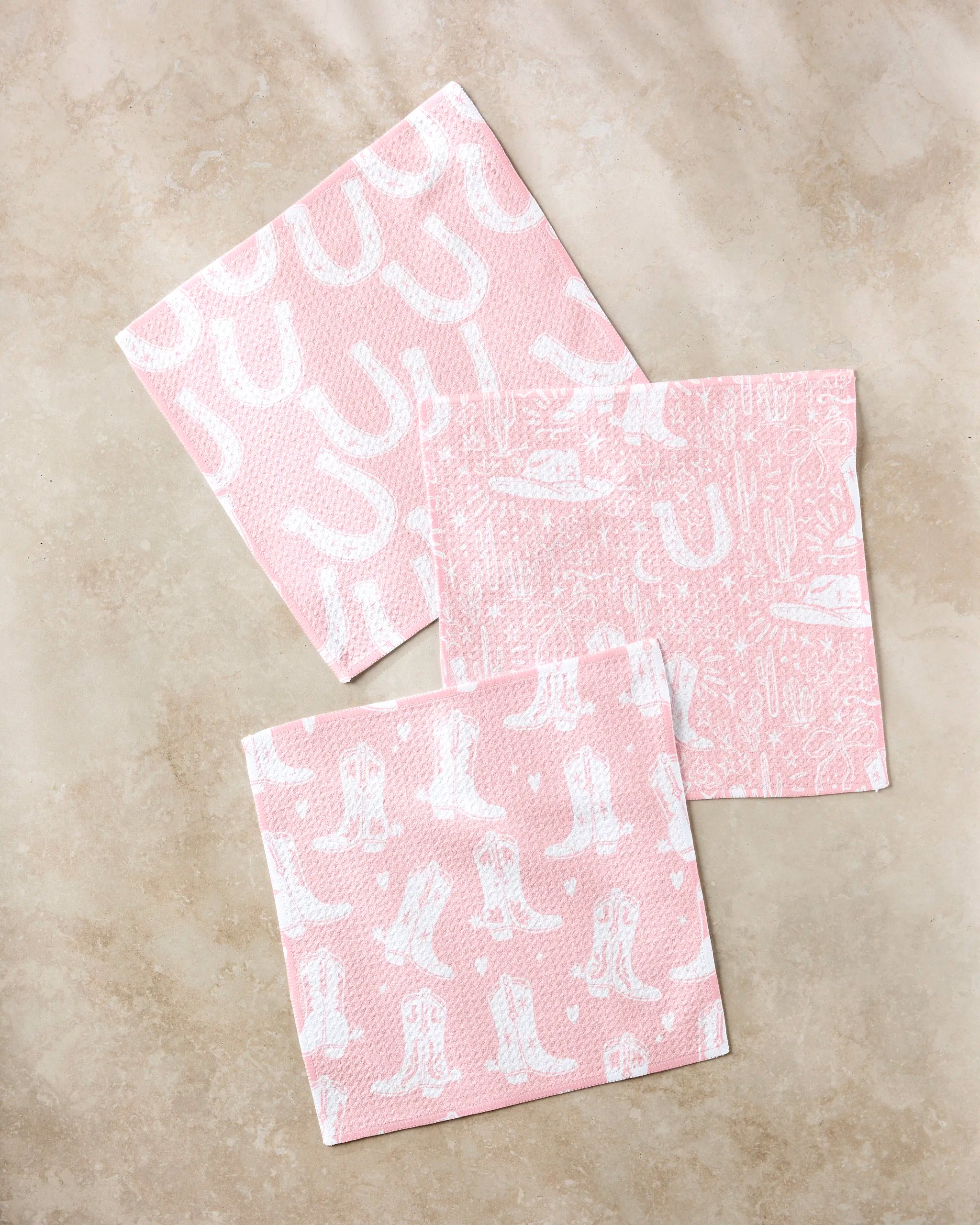 Three Aubrey Fairchild Chic Lucky Boots pink napkins, featuring white Western motifs—cowboy boots, hats, and horseshoes—lie on beige marble; perfect to match with an absorbent dishcloth set for your next themed party.