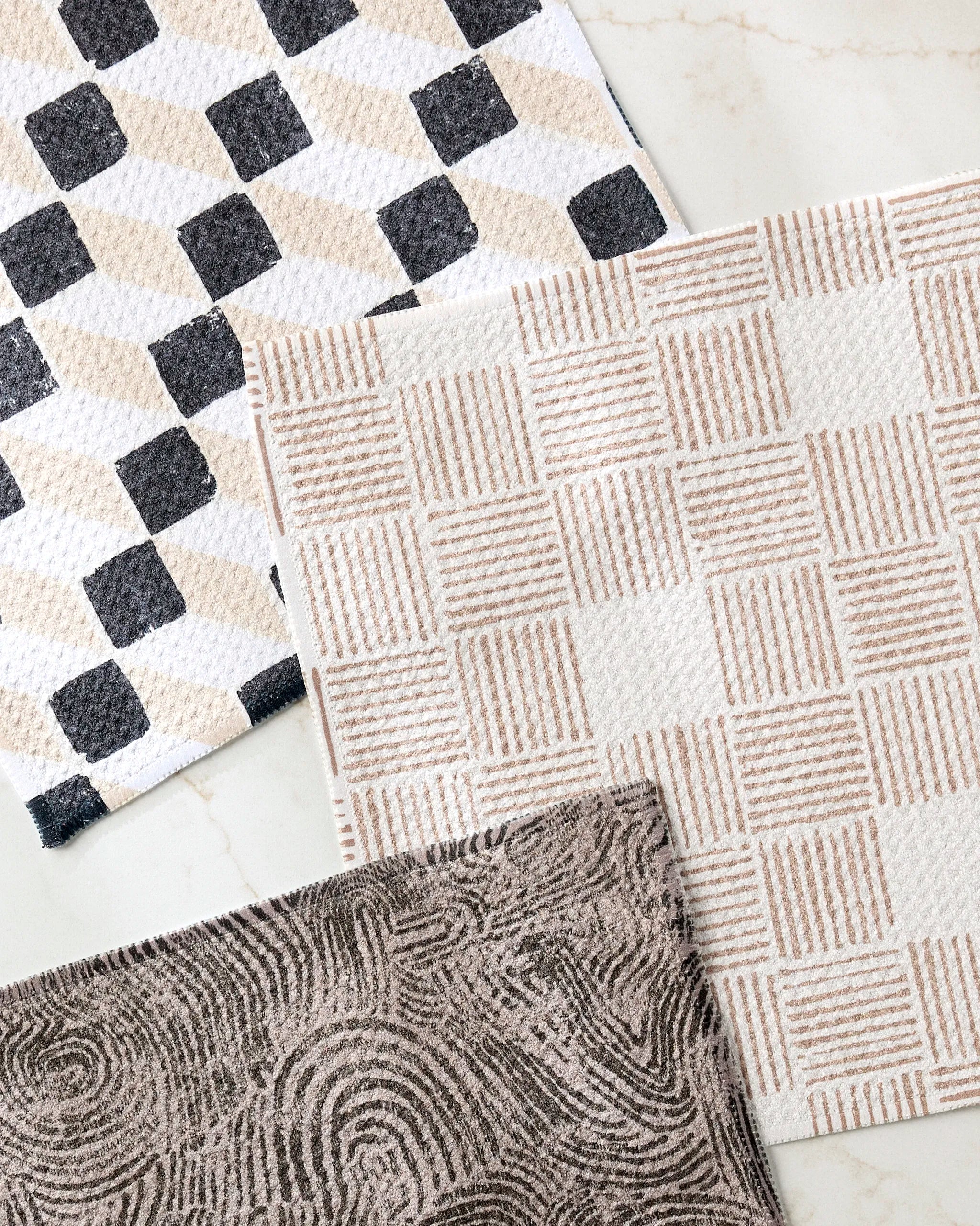 Three Chiseled mats by Studio Jeannot overlap on a light surface: one with a black-beige check pattern, one cream-tan grid, and one brown with swirls—each made from recycled materials for stylish, eco-friendly decor.