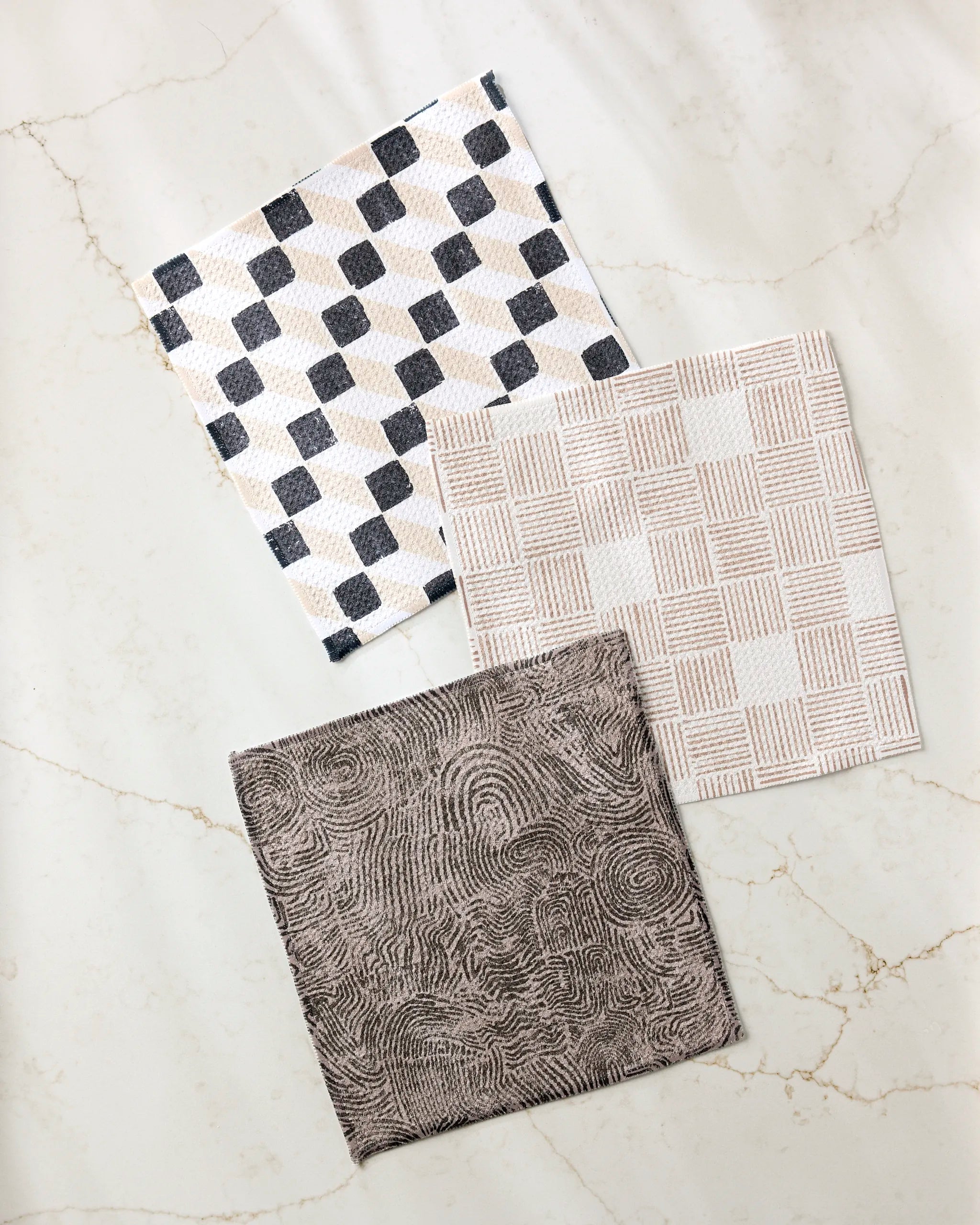 Studio Jeannot’s Chiseled set includes three square napkins crafted from recycled materials, each with a unique geometric pattern—black and beige diamonds, beige and white squares, and brown swirls—on a marble background.
