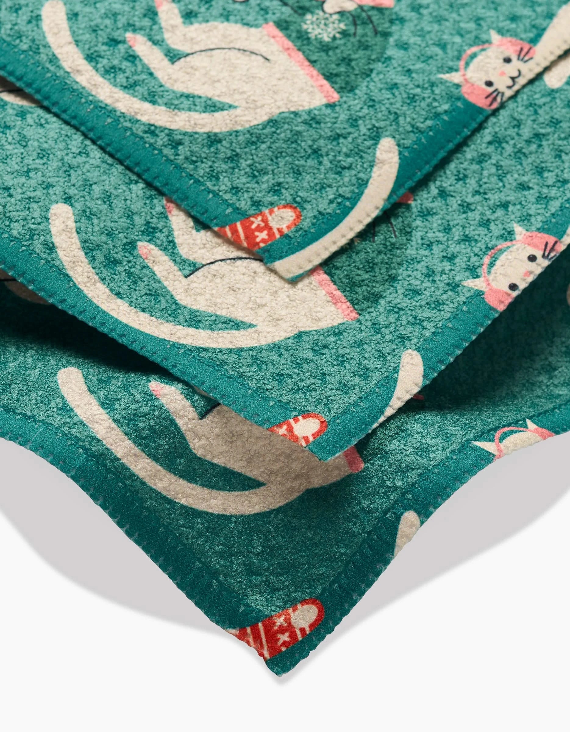 A close-up of Megan Kampas Christmas Cats dishcloths shows teal Geoweave fabric with white cats in pink hats and red scarves. Neatly stitched edges and a repeating festive cat pattern make these cloths both durable and stylish.