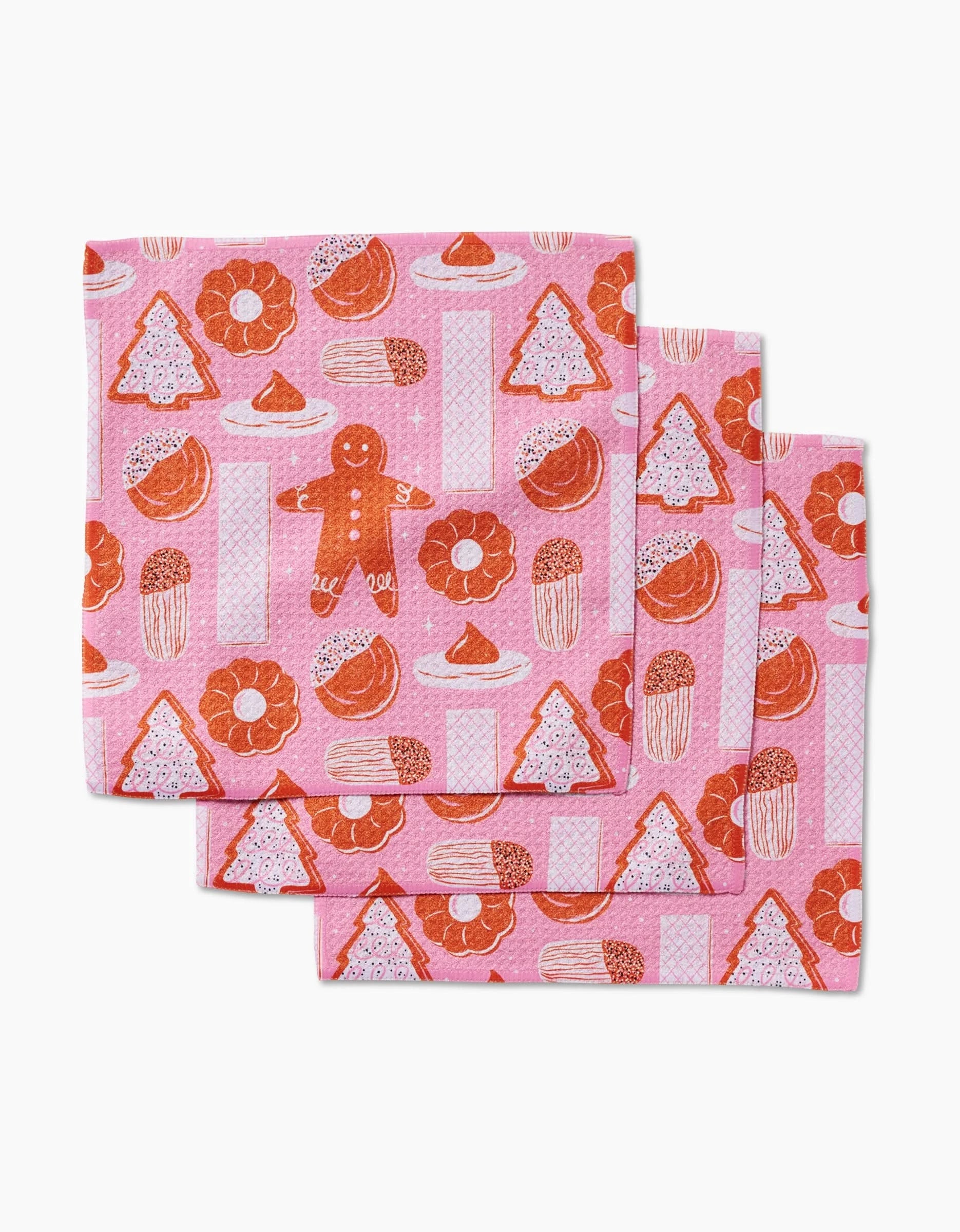Set of three folded pink napkins by Megan Roy, named Christmas Cookies, made from recycled materials and decorated with a repeating pattern of festive gingerbread men, tree-shaped cookies, and sprinkle-topped round treats.