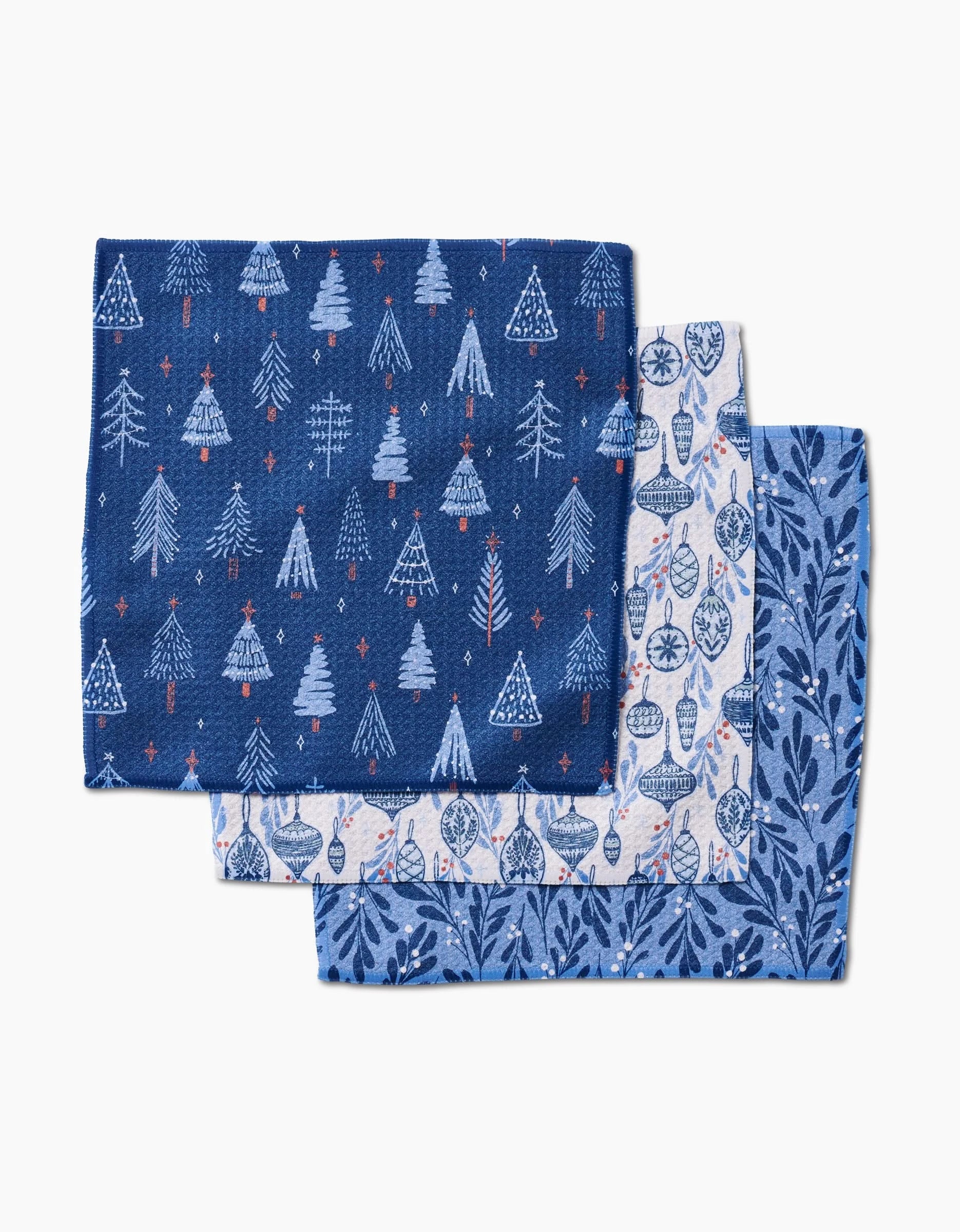 Three folded blue cloth napkins from Hufton Studio’s Christmas Decorations line, featuring winter-themed patterns—white trees and stars, ornaments, and leafy vines—are laid flat and slightly overlapping on a white background.