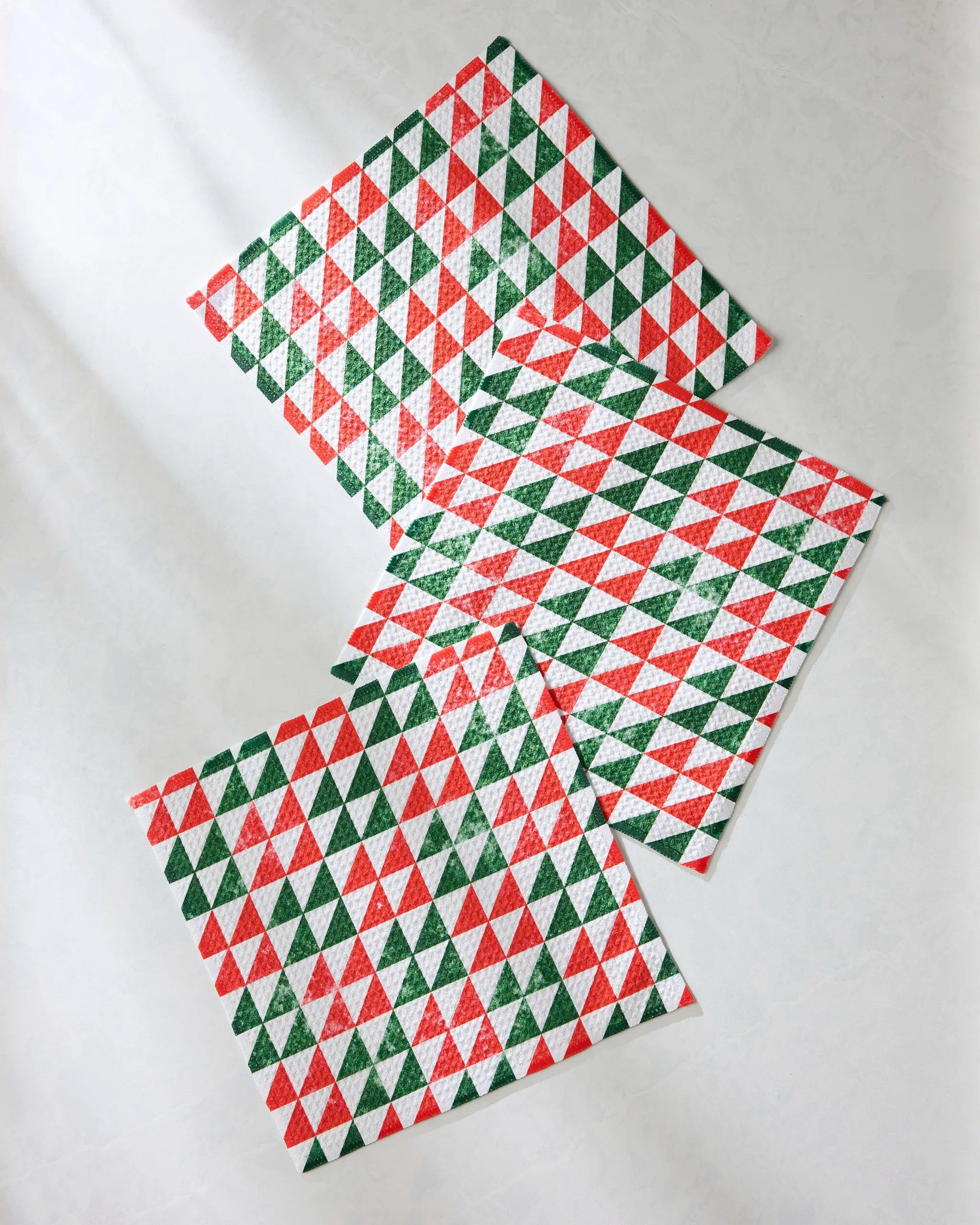 Three absorbent Geometry dishtowels from the Christmas Evergreen collection, featuring a geometric pattern of red, green, and white triangles, are arranged overlapping on a light gray surface.