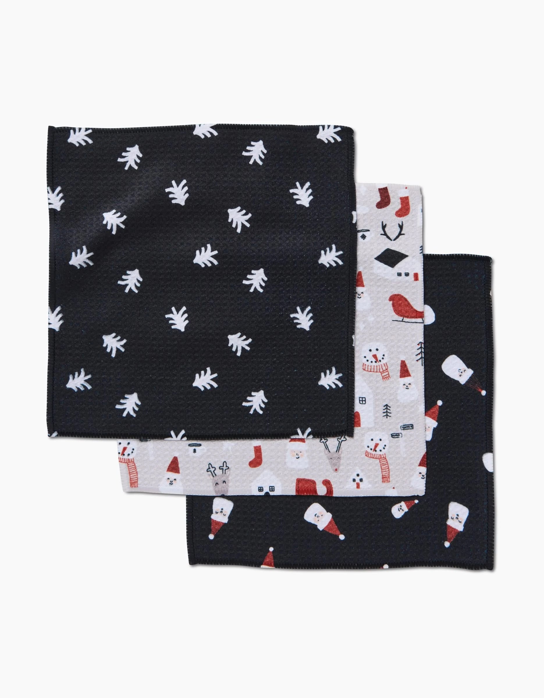 The Coit Creative Christmas Village set includes three Geoweave napkins: black with white leaves, light gray with Christmas icons, and black featuring Santa faces, snowmen, stockings, and ornaments—perfect for festive gatherings.