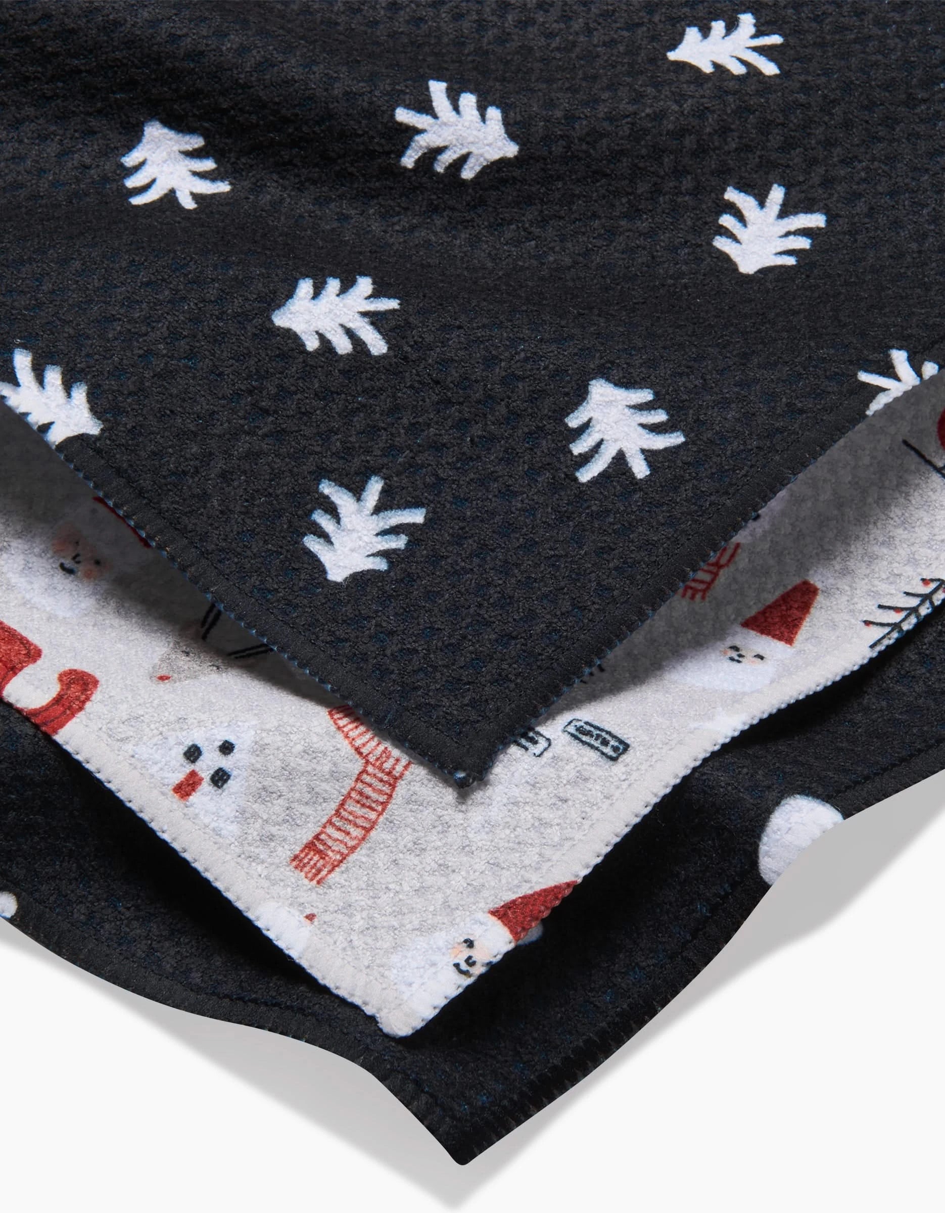 A close-up of a folded blanket shows two patterns: the top is black with white leaf prints, while the underside features Coit Creatives Christmas Village design with red and black snowmen, houses, and festive motifs.