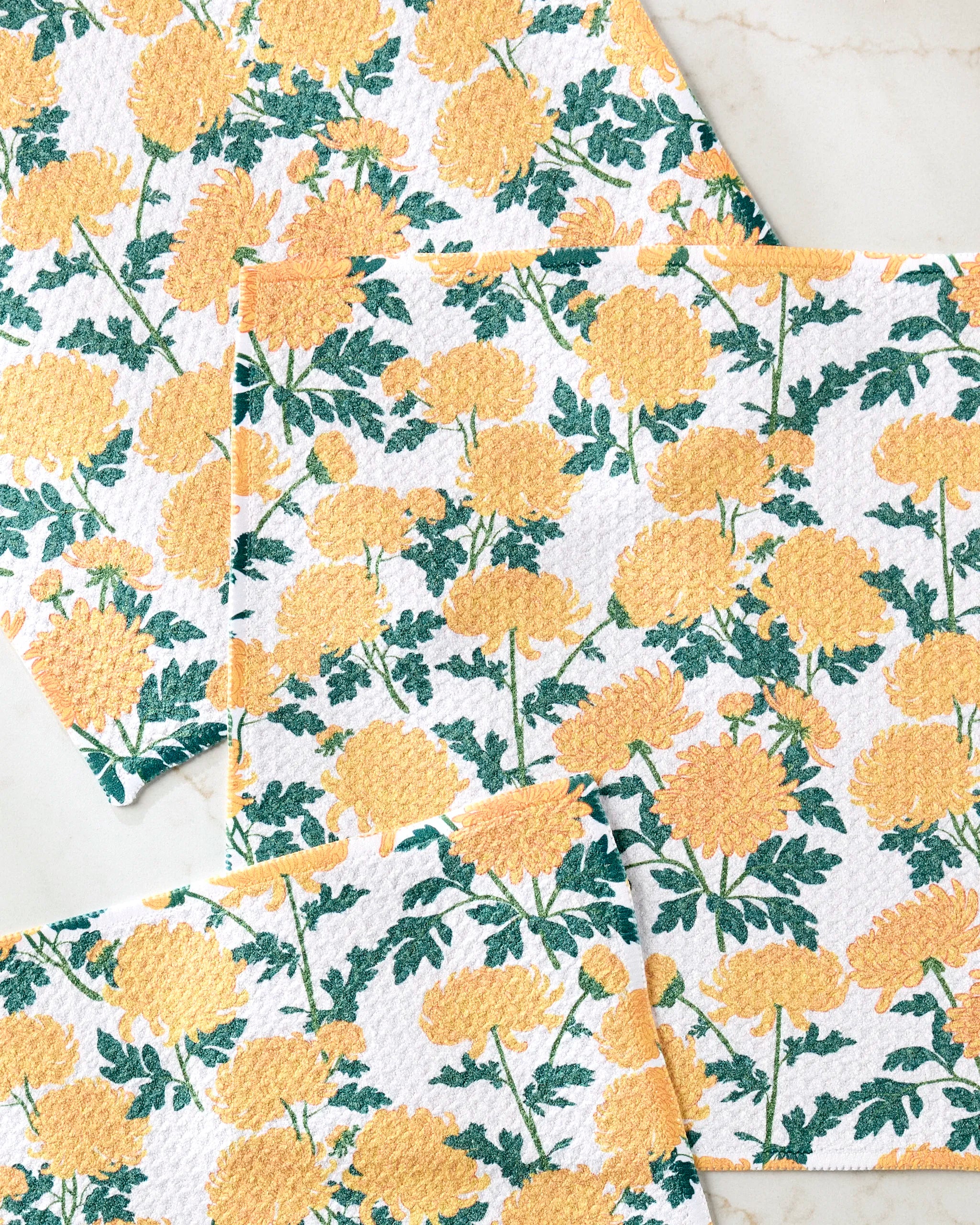 Four "Chrysanthemum" cloths by Vaishali Design Studio, made from recycled materials and featuring yellow floral and green leaf patterns, are arranged on a light, marble-style surface.