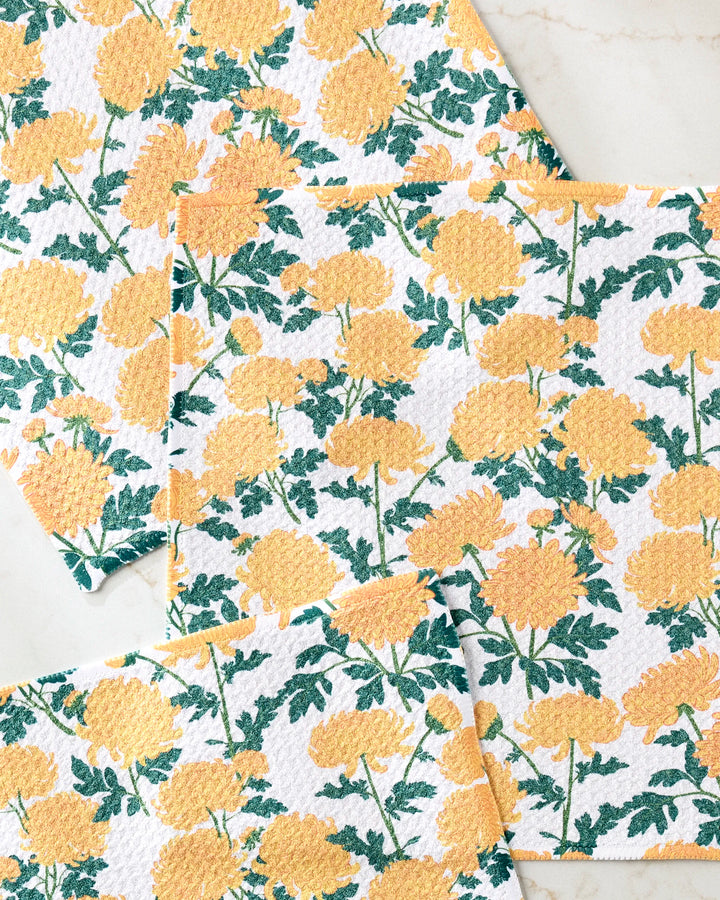 Four "Chrysanthemum" cloths by Vaishali Design Studio, made from recycled materials and featuring yellow floral and green leaf patterns, are arranged on a light, marble-style surface.