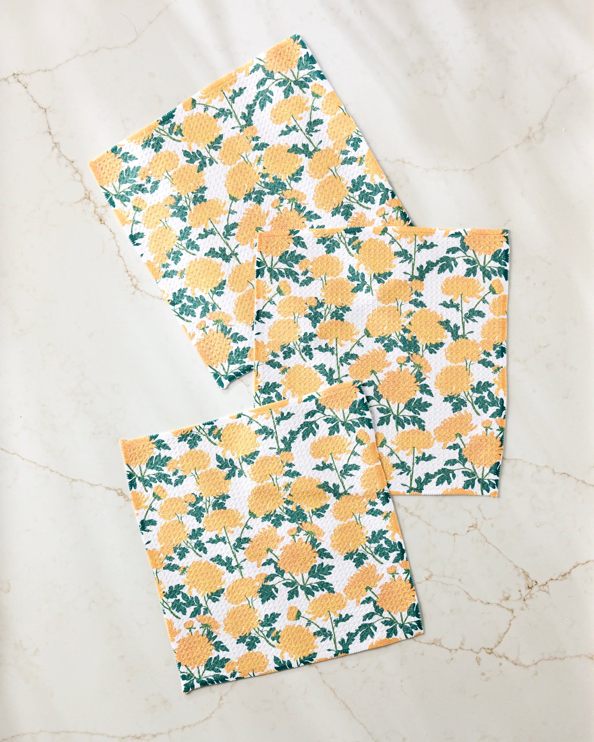 Three "Chrysanthemum" square paper napkins by Vaishali Design Studio, featuring a yellow floral and green leaf pattern, are arranged diagonally on marble—an ideal match for any eco-friendly dishcloth set.