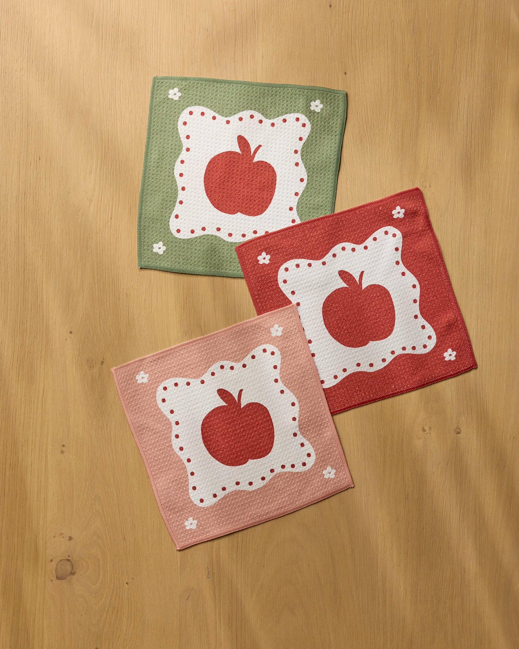Three square Geometry Cider Mill kitchen towels with apple motifs, white floral accents, and decorative borders in green, red, and pink hues are displayed on a wooden surface. Each eco-friendly dishcloth is made from recycled materials.