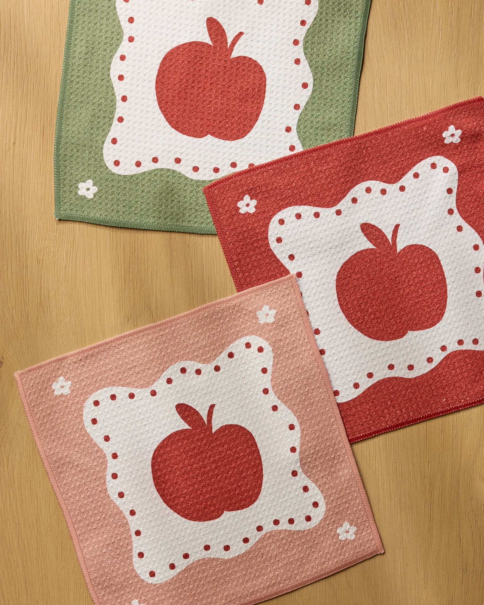 Three square Cider Mill kitchen towels by Geometry, in green, red, and pink, each display a red apple with white flower accents in the corners and are arranged on a wooden surface.