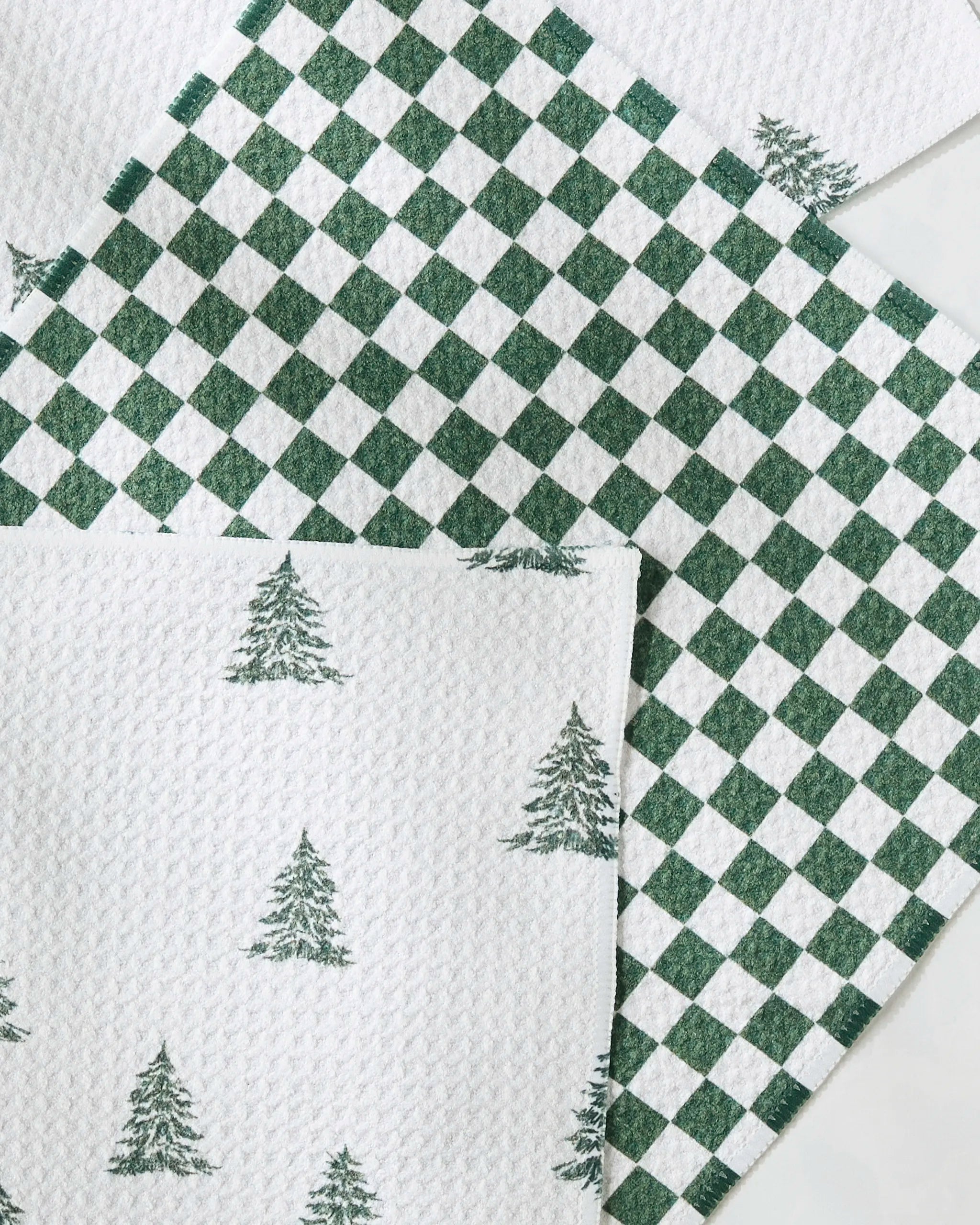 Three textured, absorbent Claret kitchen towels by Geometry overlap—two with green Christmas tree prints on white and one with a green and white checkered pattern—creating a festive, holiday-themed dishcloth set.