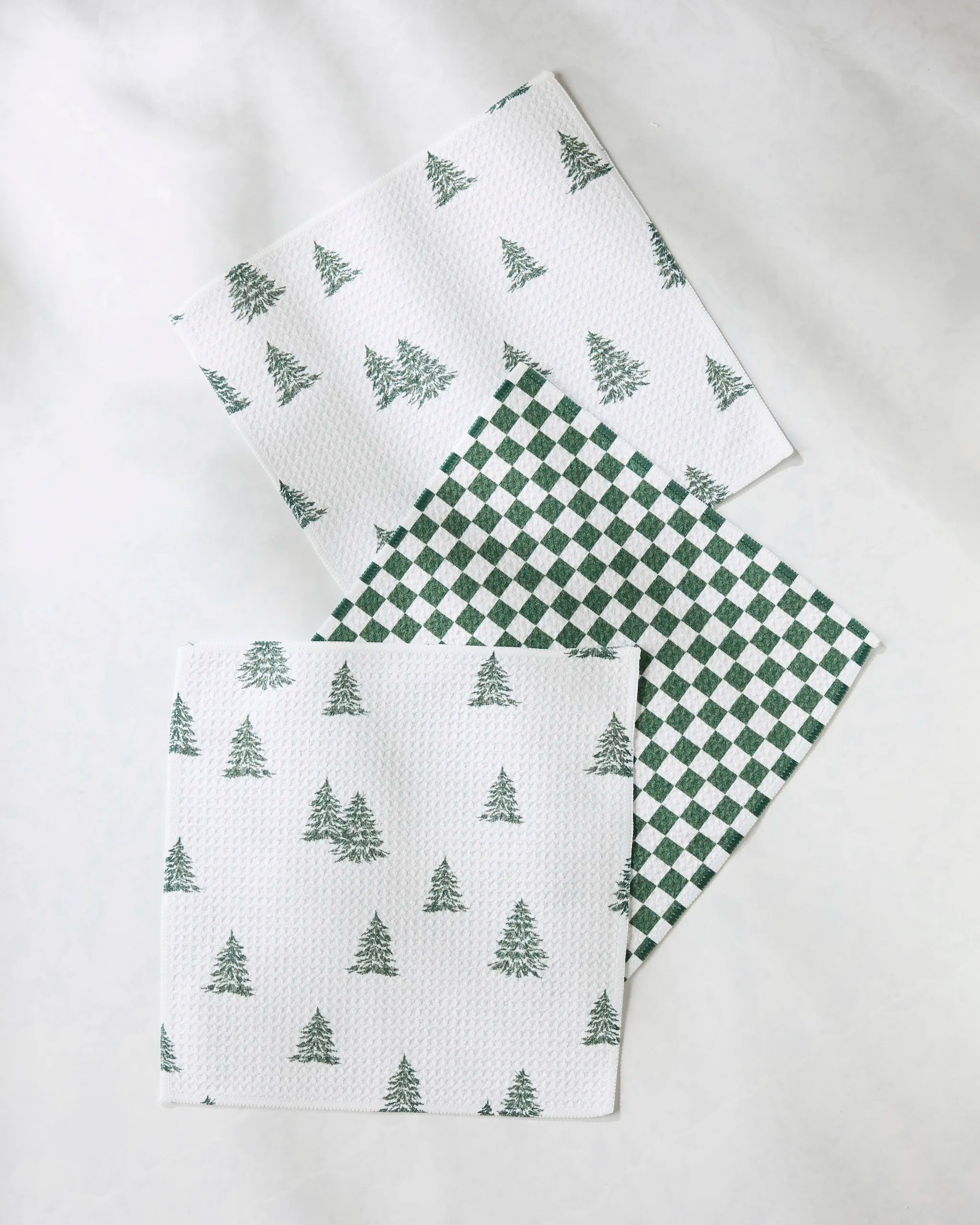 The Claret set by Geometry includes three eco-friendly recycled dishcloths: two with a green pine tree pattern and one with a green and white checkered design—perfect for your kitchen.