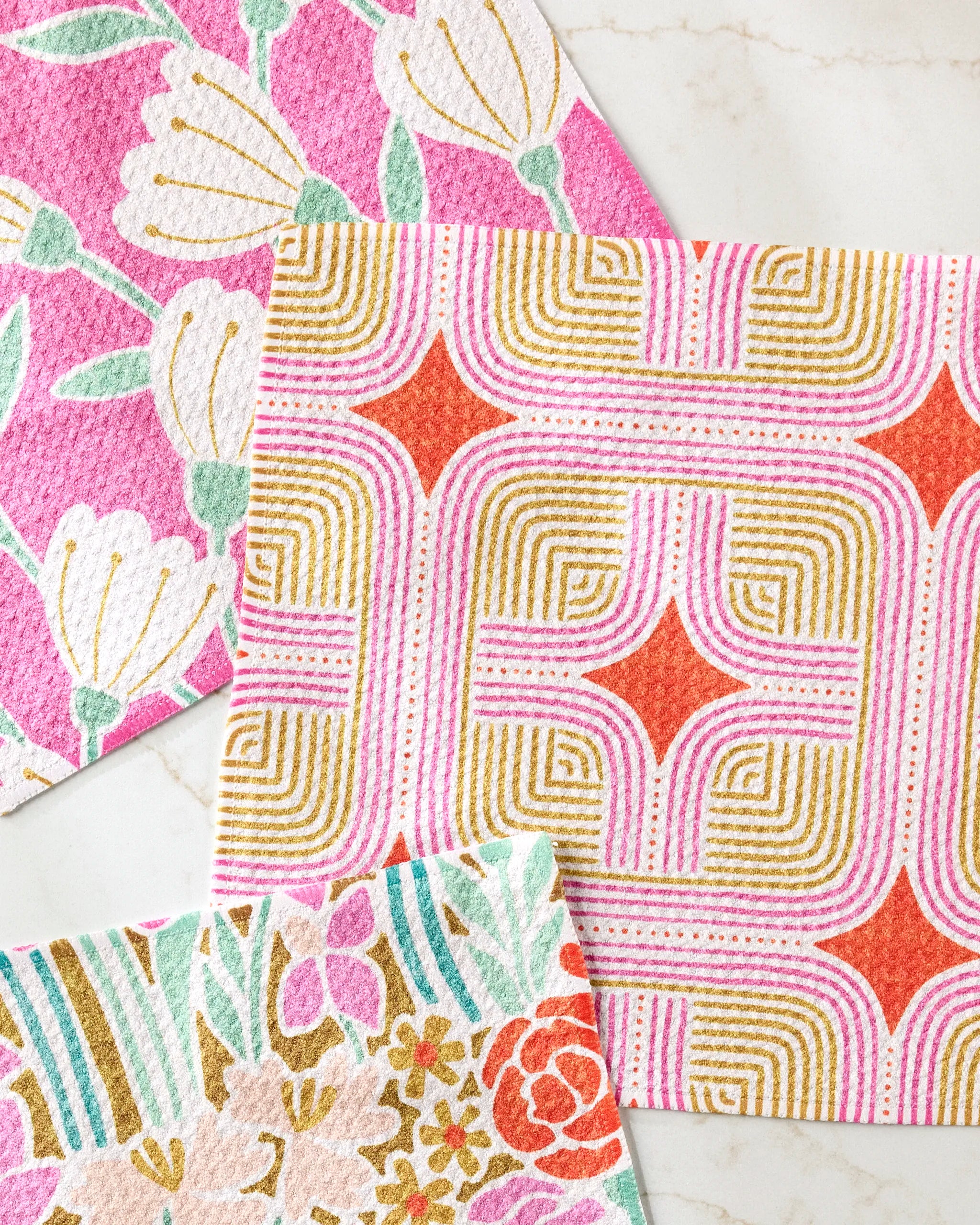 Three colorful Clemmie Donna and Janae dishcloths by Amy MacCready are displayed—one with pink and white flowers, one with geometric orange, pink, and gold shapes, and another featuring pastel floral patterns.