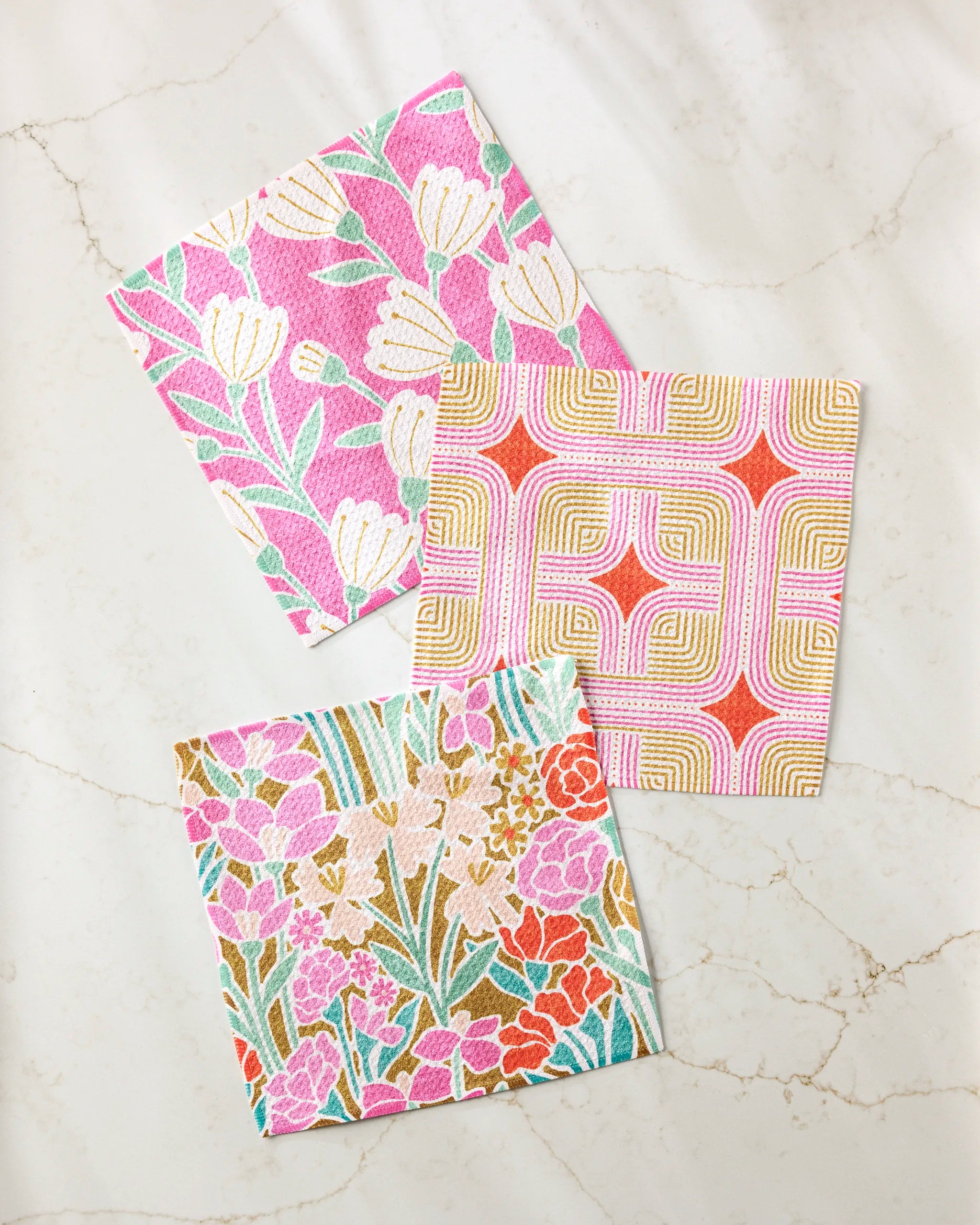 Three colorful square napkins from Amy MacCready's Clemmie Donna and Janae set, featuring floral and geometric motifs in pink, orange, green, and gold tones, are arranged on a light marble surface.