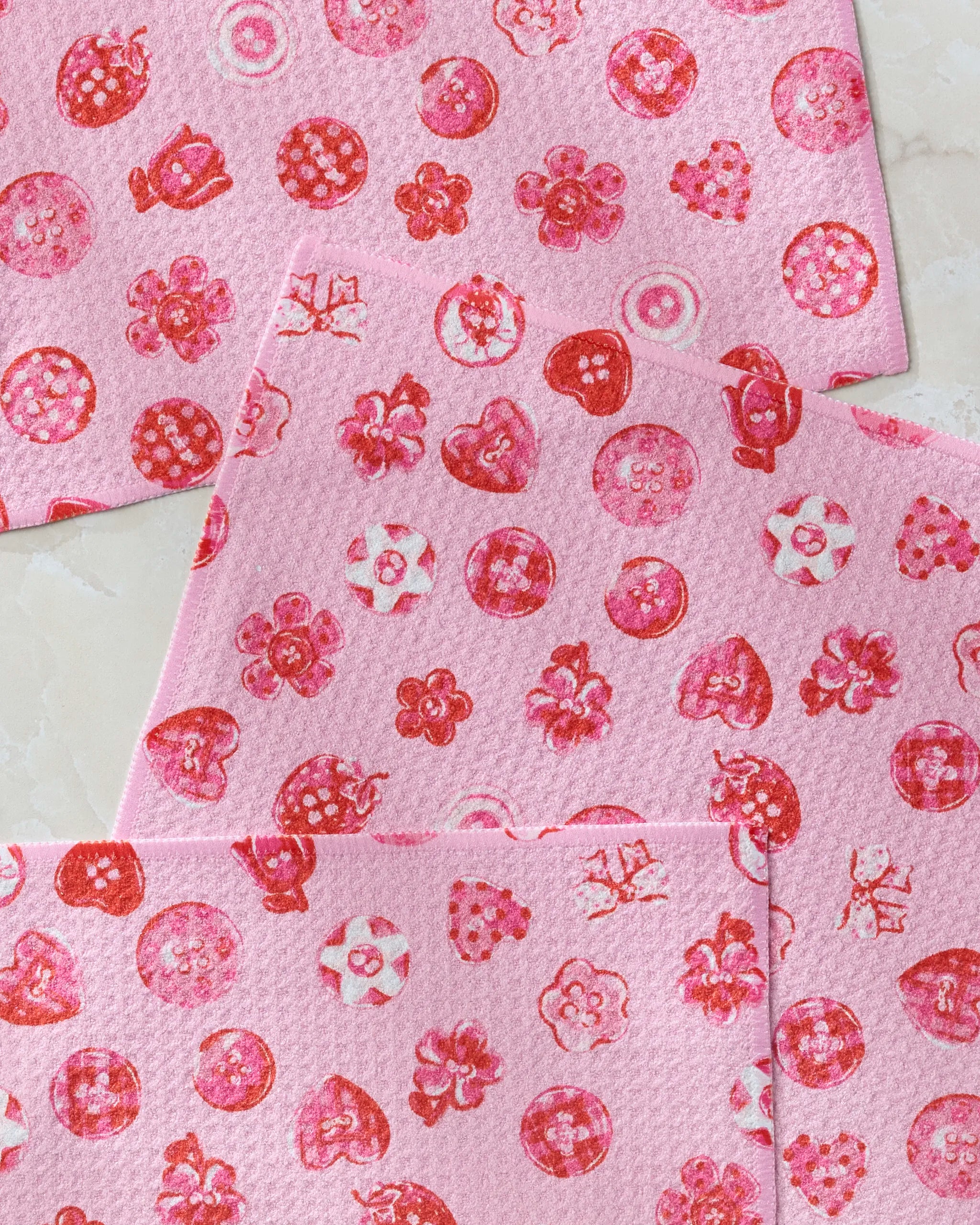 Three pink Collecting Buttons dish towels by Geometry, featuring red and white floral and geometric patterns with flowers, circles, and decorative shapes, are layered on a light-colored surface. Eco-friendly design.