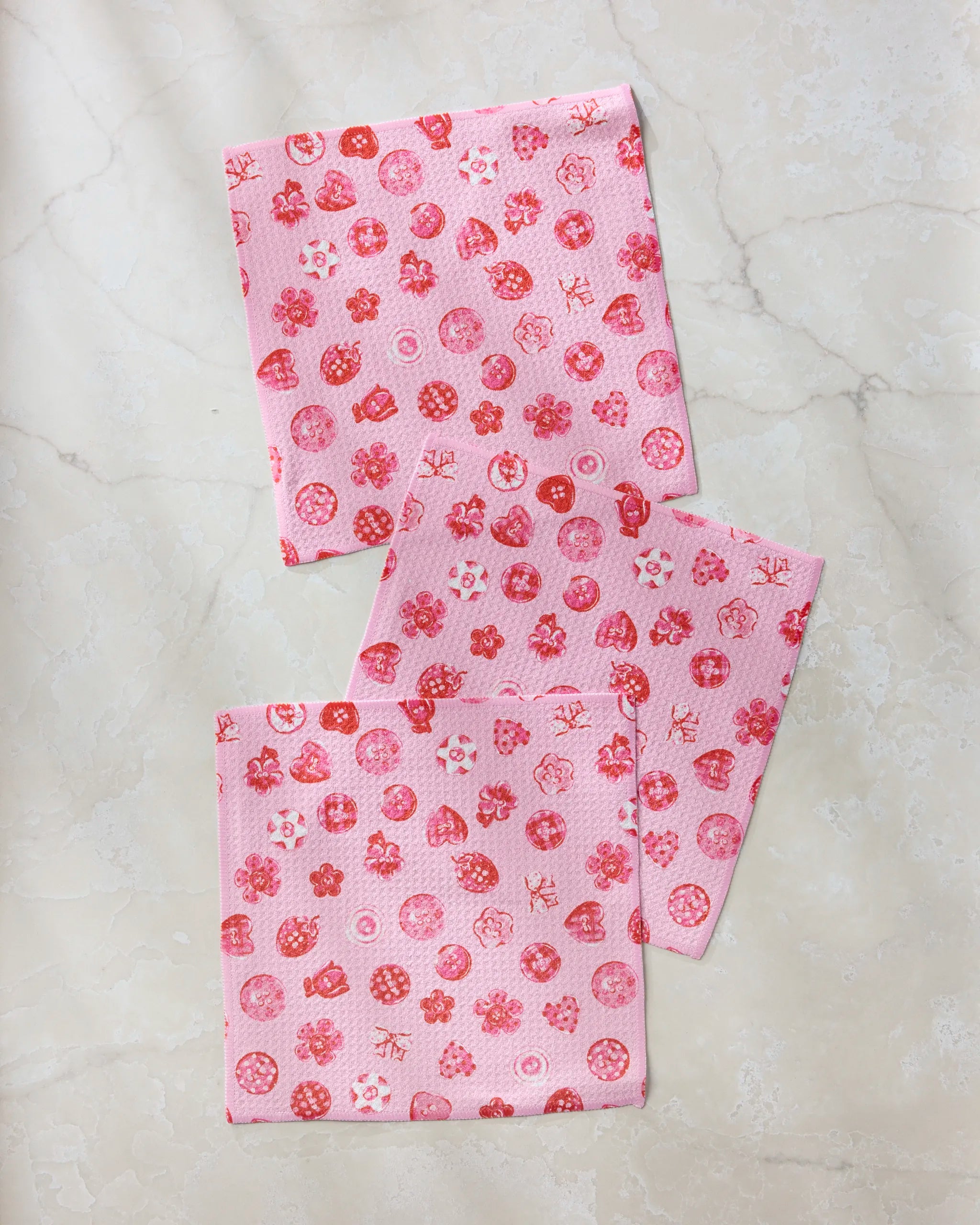 The Geometry Collecting Buttons set features three pink square eco-friendly dishcloths with red and white floral and geometric patterns, stylishly arranged on a light, textured surface for an elegant kitchen display.