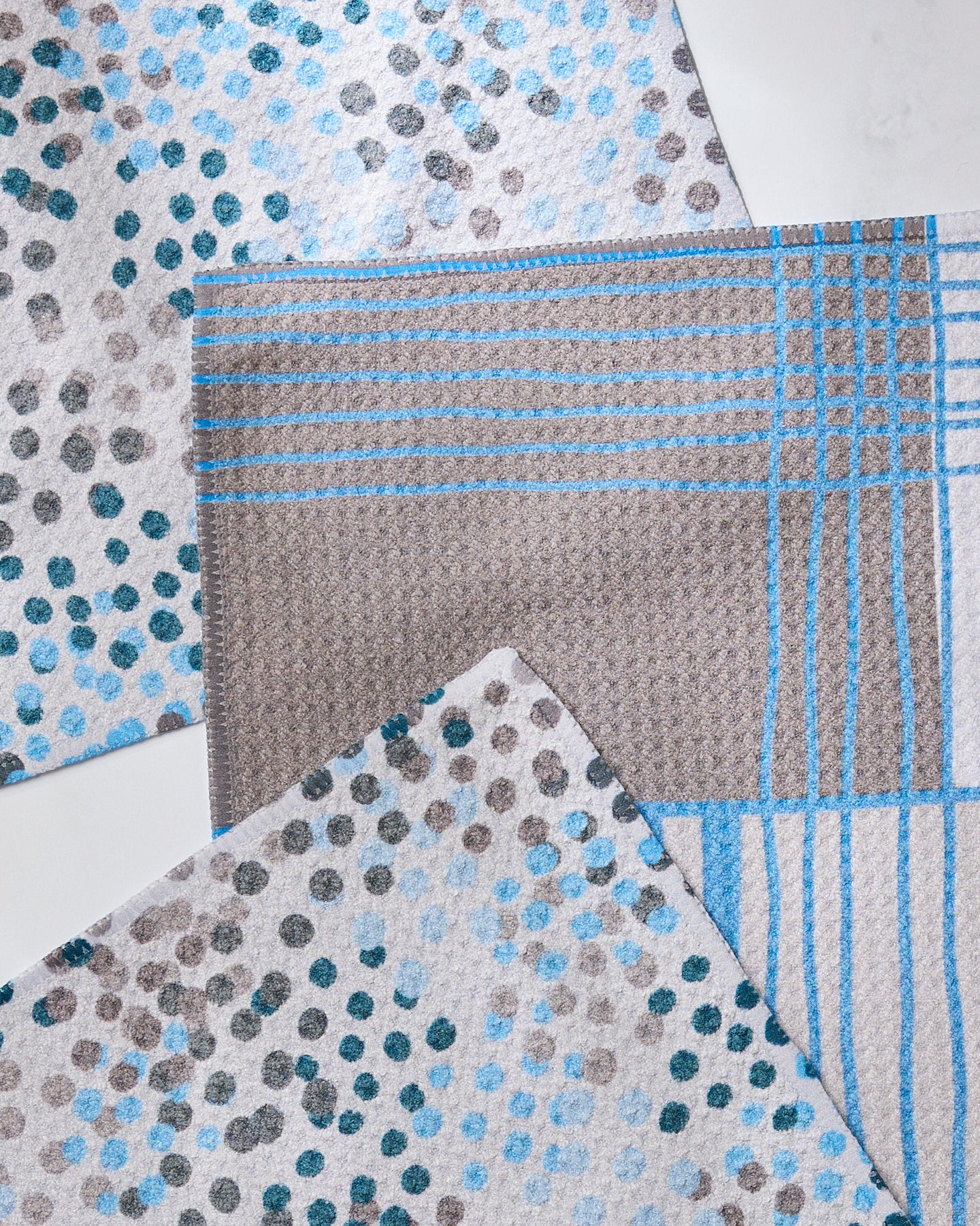 Three Cool Blocks and Dots fabric samples by Geometry are neatly arranged on a white surface, showcasing blue, gray, and white recycled materials—two with scattered dots and one featuring a grid of blue lines on gray.